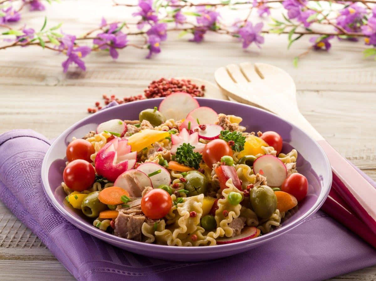 Healthy Food 桌布 壁紙 — 一碗充滿活力的義大利麵沙拉，配有櫻桃番茄、蘿蔔、橄欖和歐芹，放在紫色碗中，置於木桌上，伴以紫色花朵。