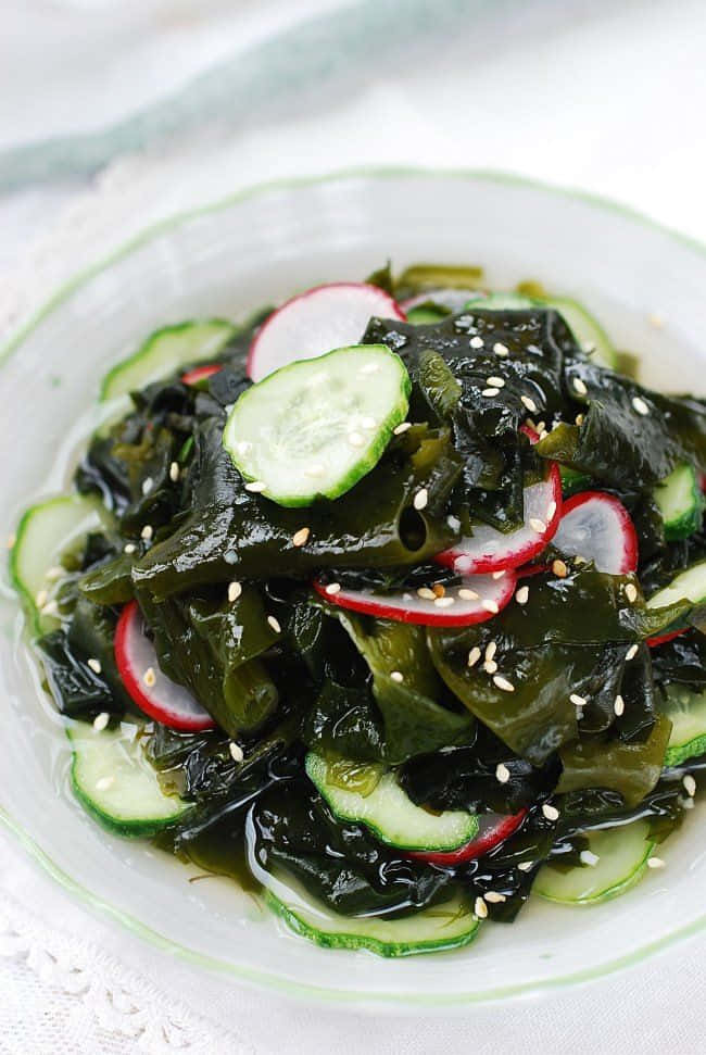 Seaweed 桌布 壁紙 — 一碗光澤的海藻沙拉，上面放有黃瓜、蘿蔔和芝麻，放在蕾絲桌布上。