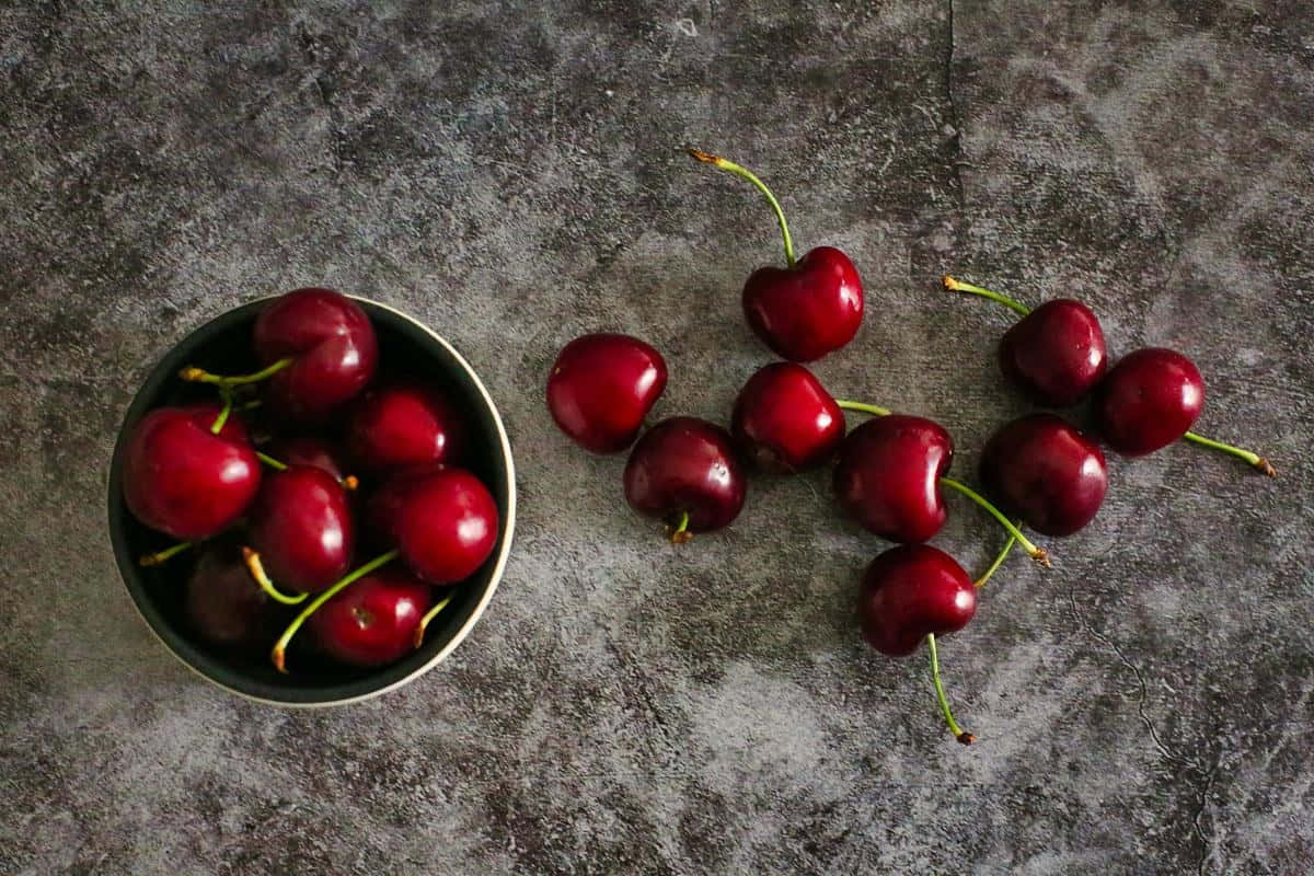 Red Cherries 桌布 壁紙 — 一碗光澤的紅櫻桃放在質感灰色石面上，旁邊散落著櫻桃。