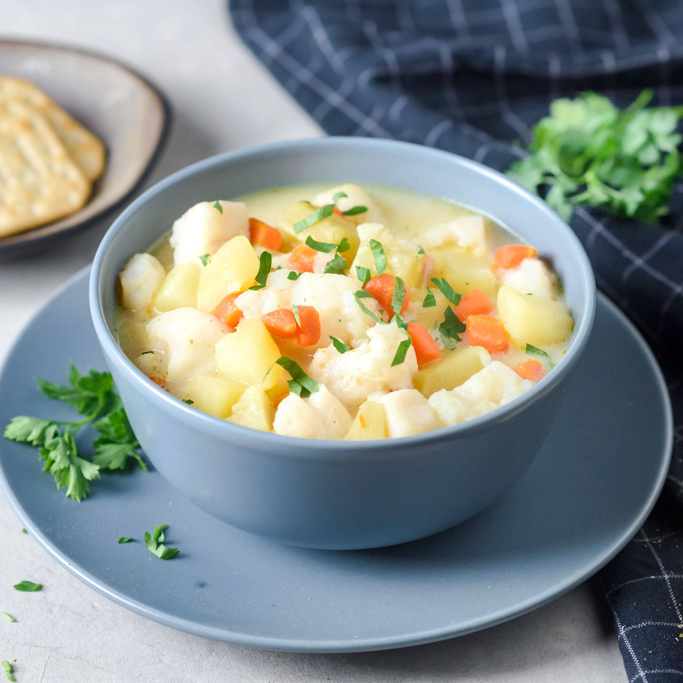 Chowder 桌布 壁紙 — 一碗冒煙的奶油魚湯，配有馬鈴薯和紅蘿蔔，上面撒上新鮮歐芹，放在灰色盤子上，旁邊有格紋餐巾和麵包。