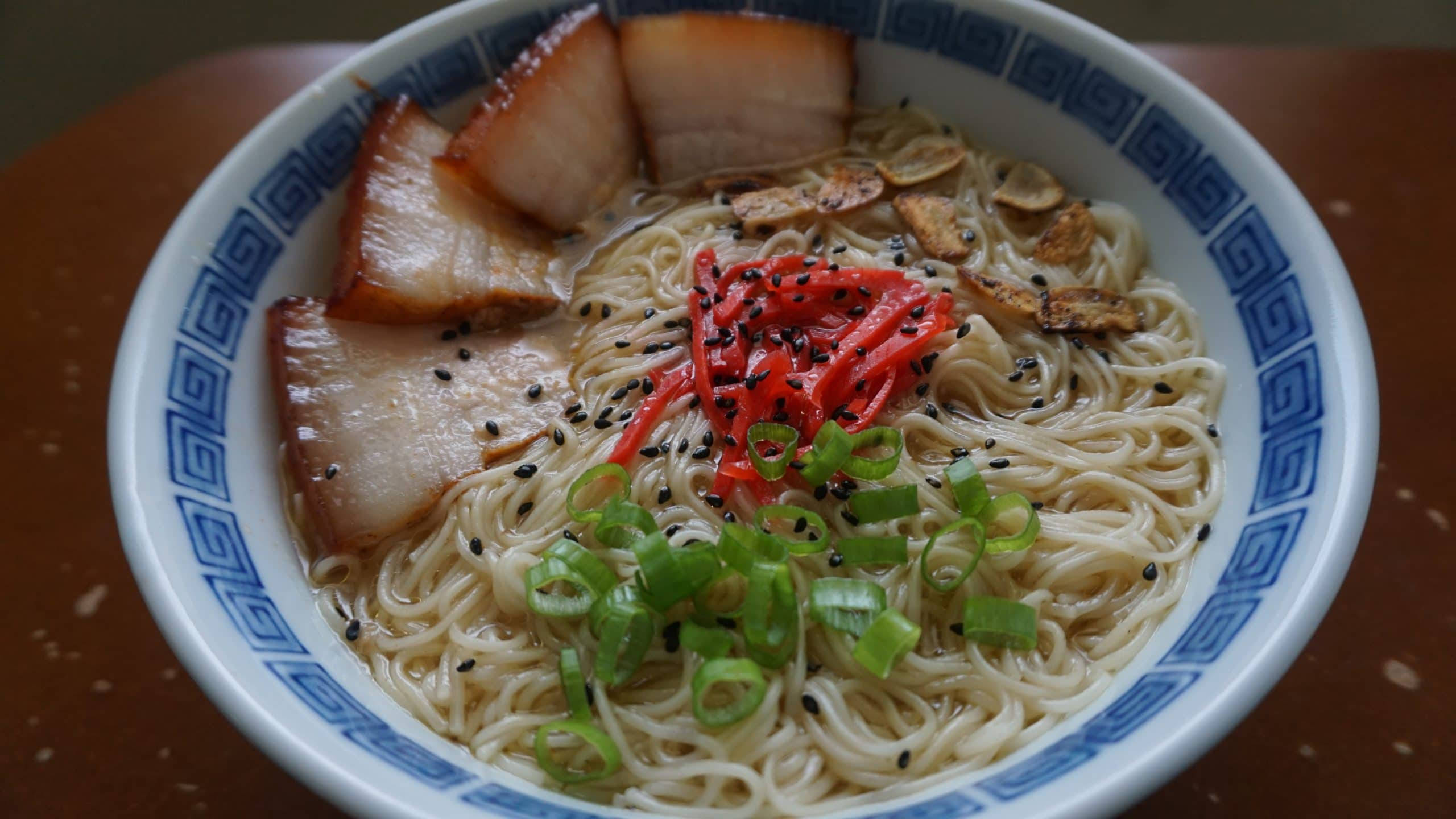 Noodles 桌布 壁紙 — 一碗冒煙的拉麵，上面鋪著脆皮豬肉片、醃薑、青蔥和黑芝麻，盛在藍色花紋碗中。