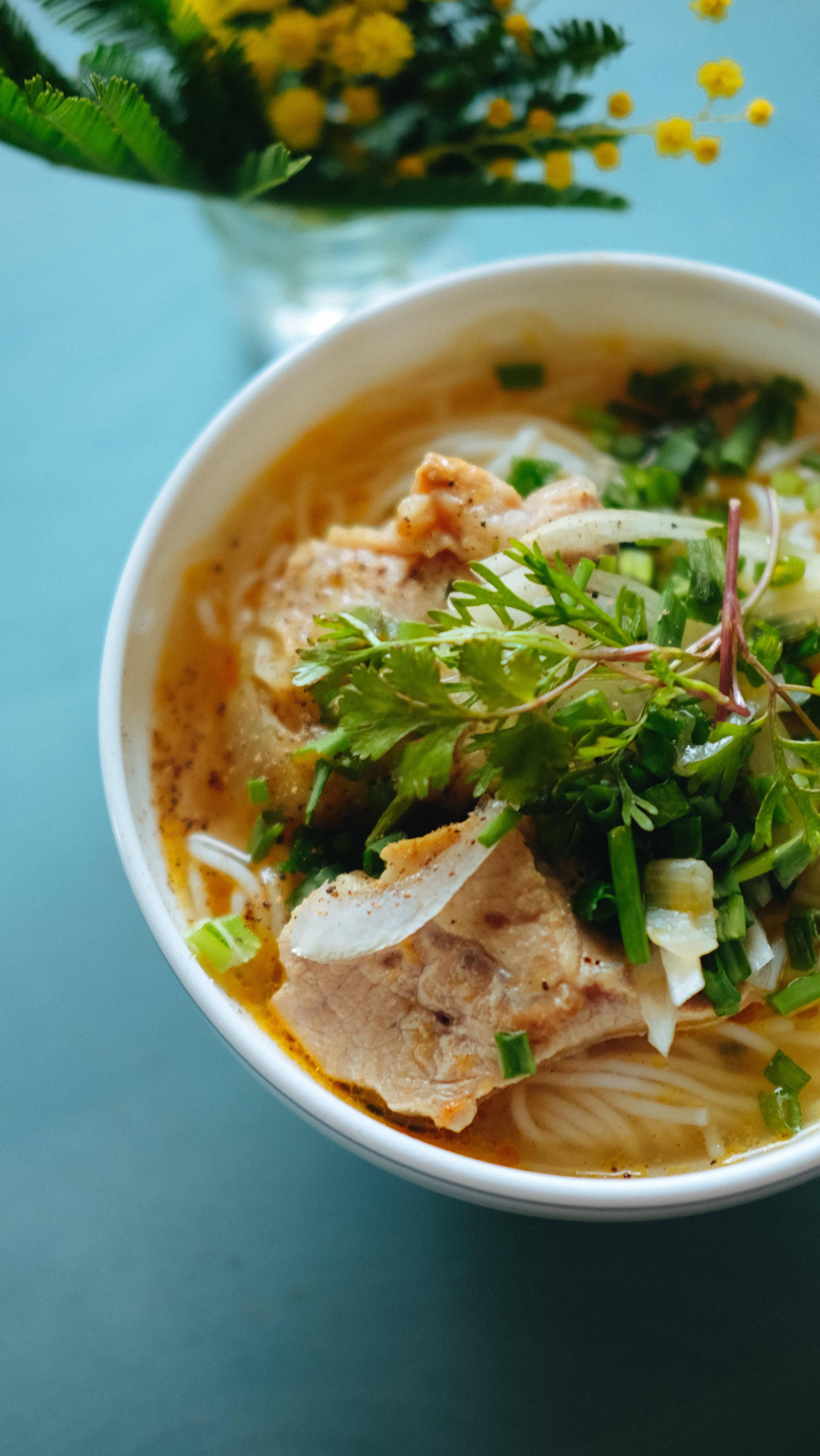 Noodles 桌布 壁紙 — 一碗冒煙的湯麵，上面鋪滿新鮮香草和切片肉，旁邊放著一瓶黃花，放在青綠色的表面上。