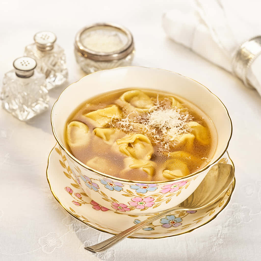 Food Photography 桌布 壁紙 — 一碗冒煙的義大利餃子湯，撒上磨碎的起司，優雅地放在花卉圖案的碟子上，旁邊有水晶鹽和胡椒罐。