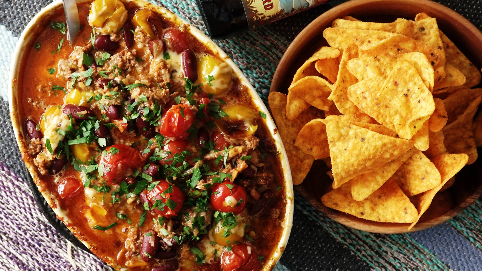 Mexican Food 桌布 壁紙 — 一碗冒煙的豐盛辣椒，上面鋪滿起司、櫻桃番茄和香菜，旁邊放著一碗木碗盛裝的調味墨西哥玉米片。