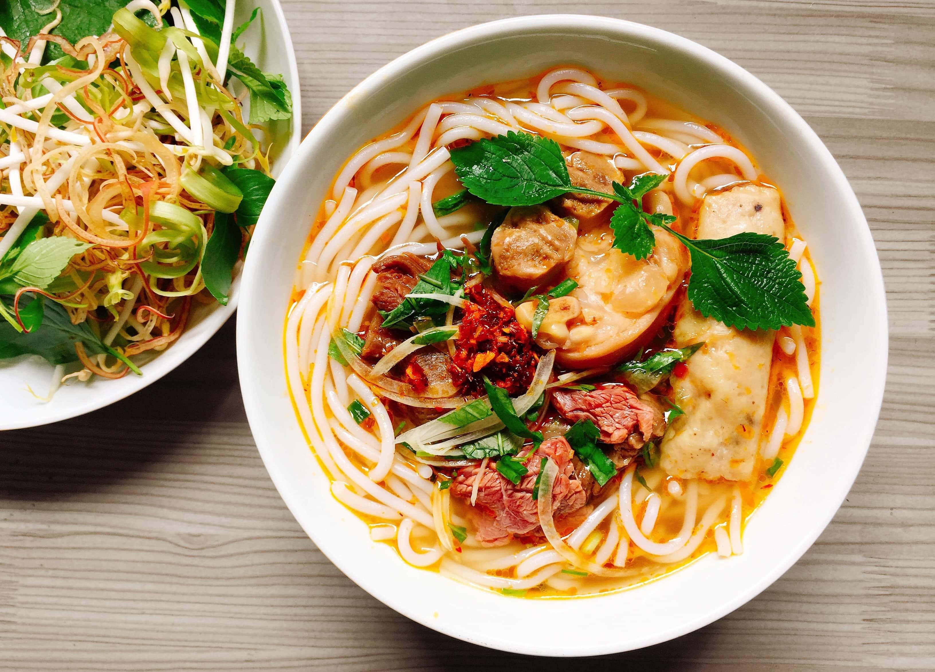 Food Photography 桌布 壁紙 — 一碗冒煙的越南河粉，配有米粉、牛肉、香草和辣椒油，旁邊放著一盤新鮮豆芽和青菜，在木桌上。