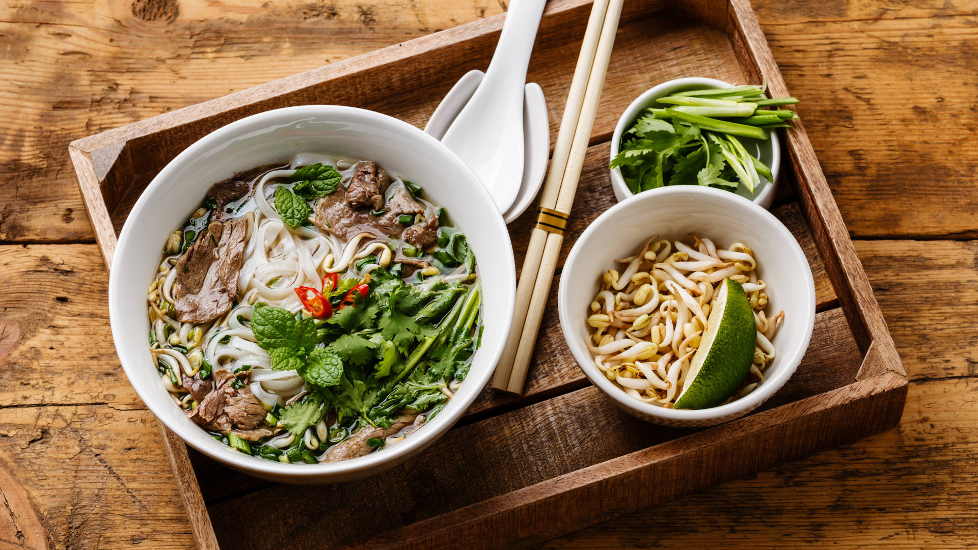 Food Photography 桌布 壁紙 — 一碗冒煙的越南河粉，配有米粉、牛肉、香草和辣椒，放在木托盤上，附有配菜和筷子。