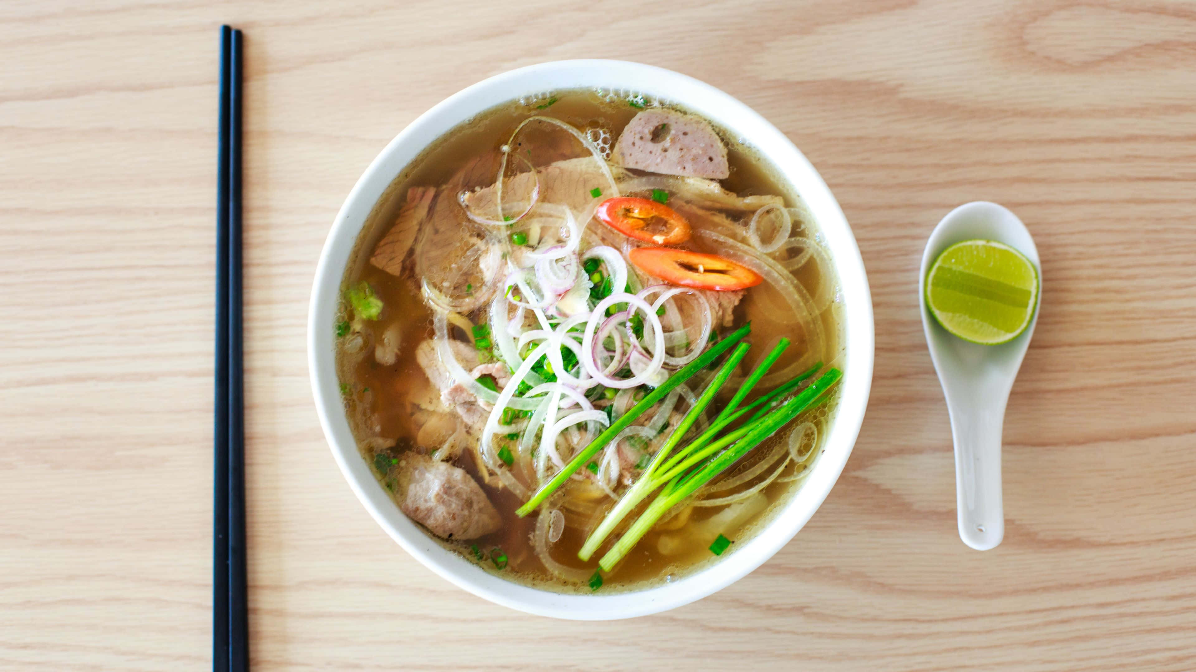 Noodles 桌布 壁紙 — 一碗冒煙的越南河粉，配有米粉、肉片、萊姆和香草，放在淺色木桌上。
