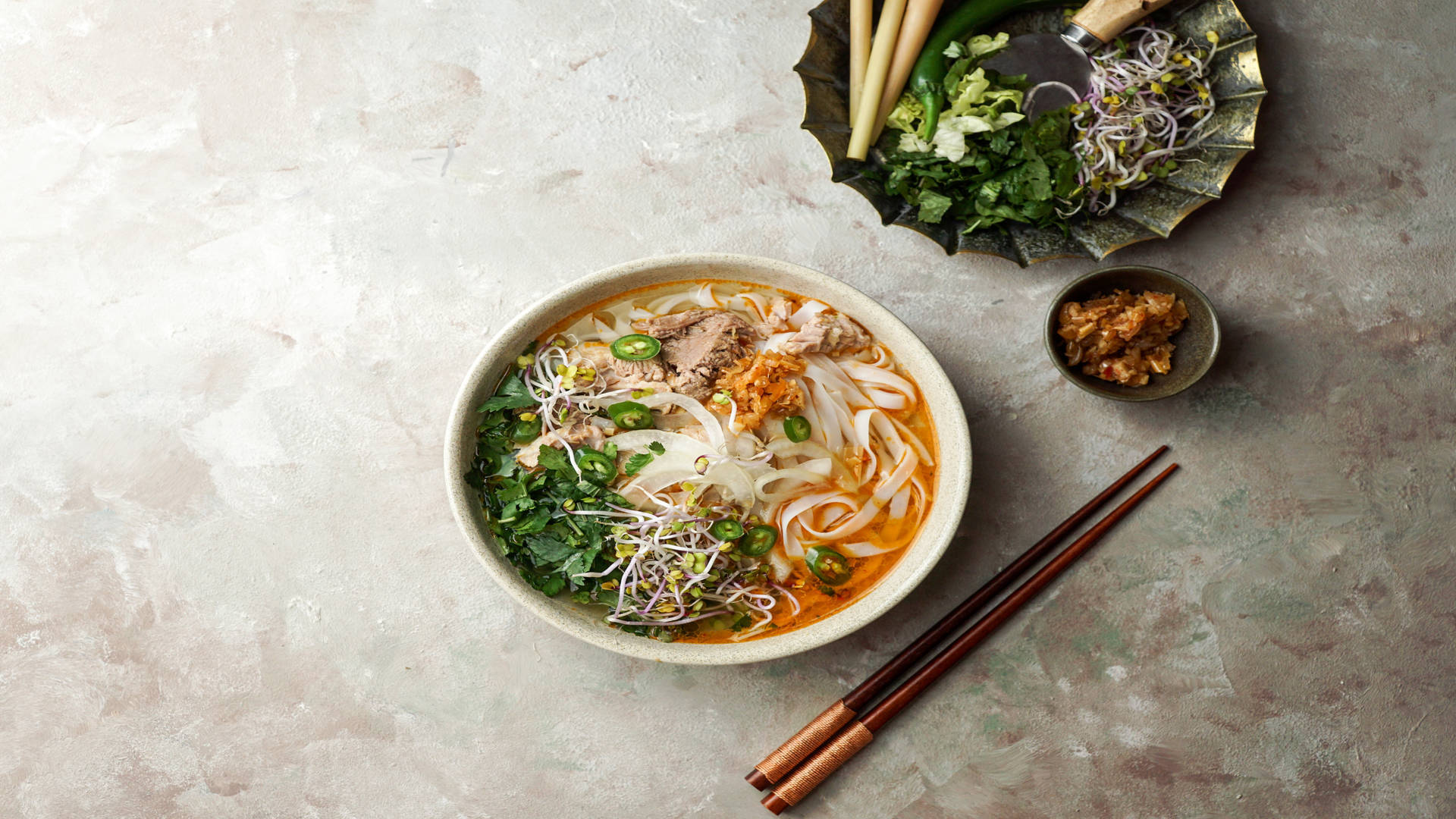 Food Photography 桌布 壁紙 — 一碗冒煙的越南河粉，配有米粉、香草和肉類，旁邊放著筷子和配菜，置於有紋理的石面上。