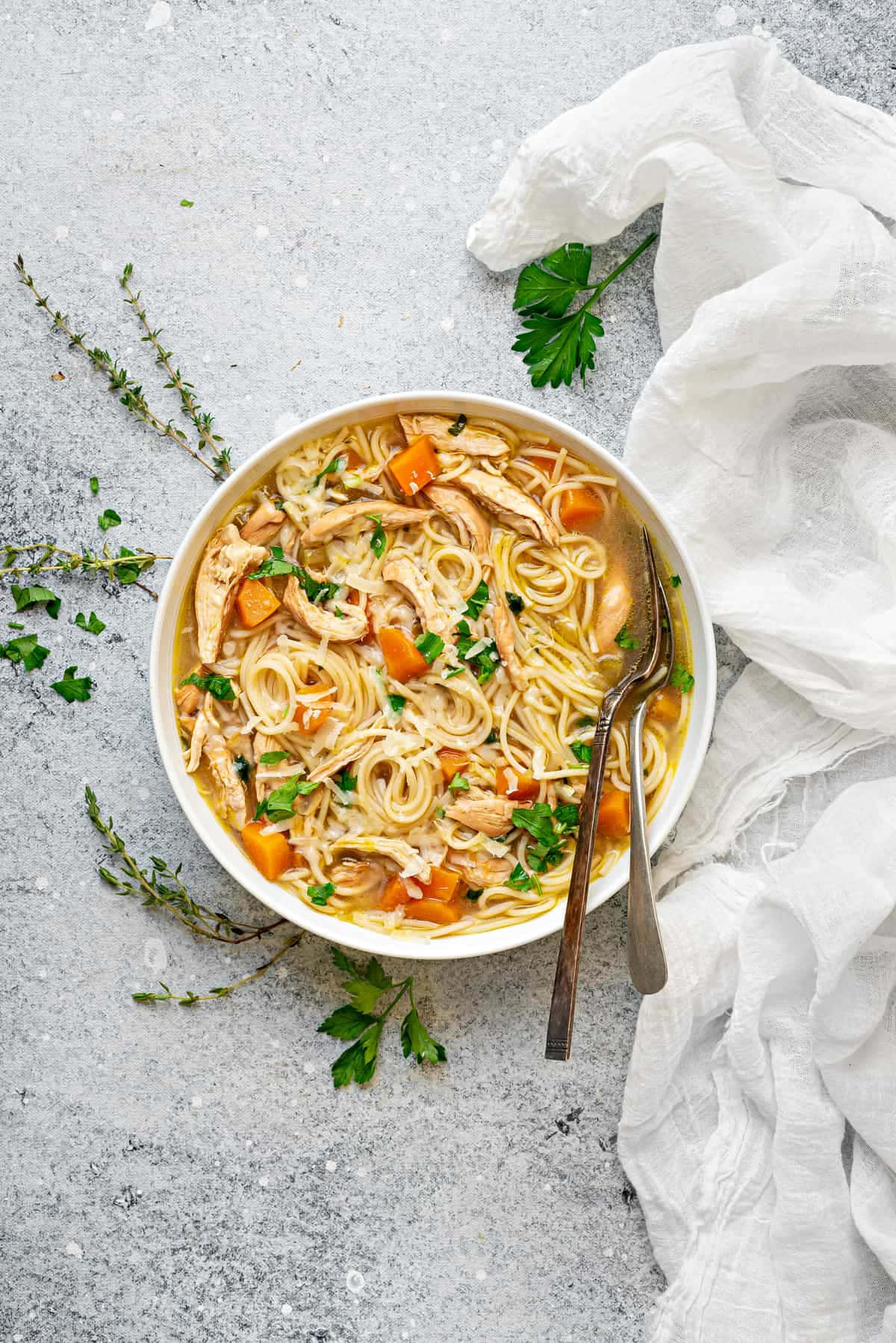 Noodles 桌布 壁紙 — 一碗冒煙的雞肉麵湯，飾以歐芹和紅蘿蔔，旁邊放著一塊白布和新鮮香草，置於有紋理的灰色表面上。