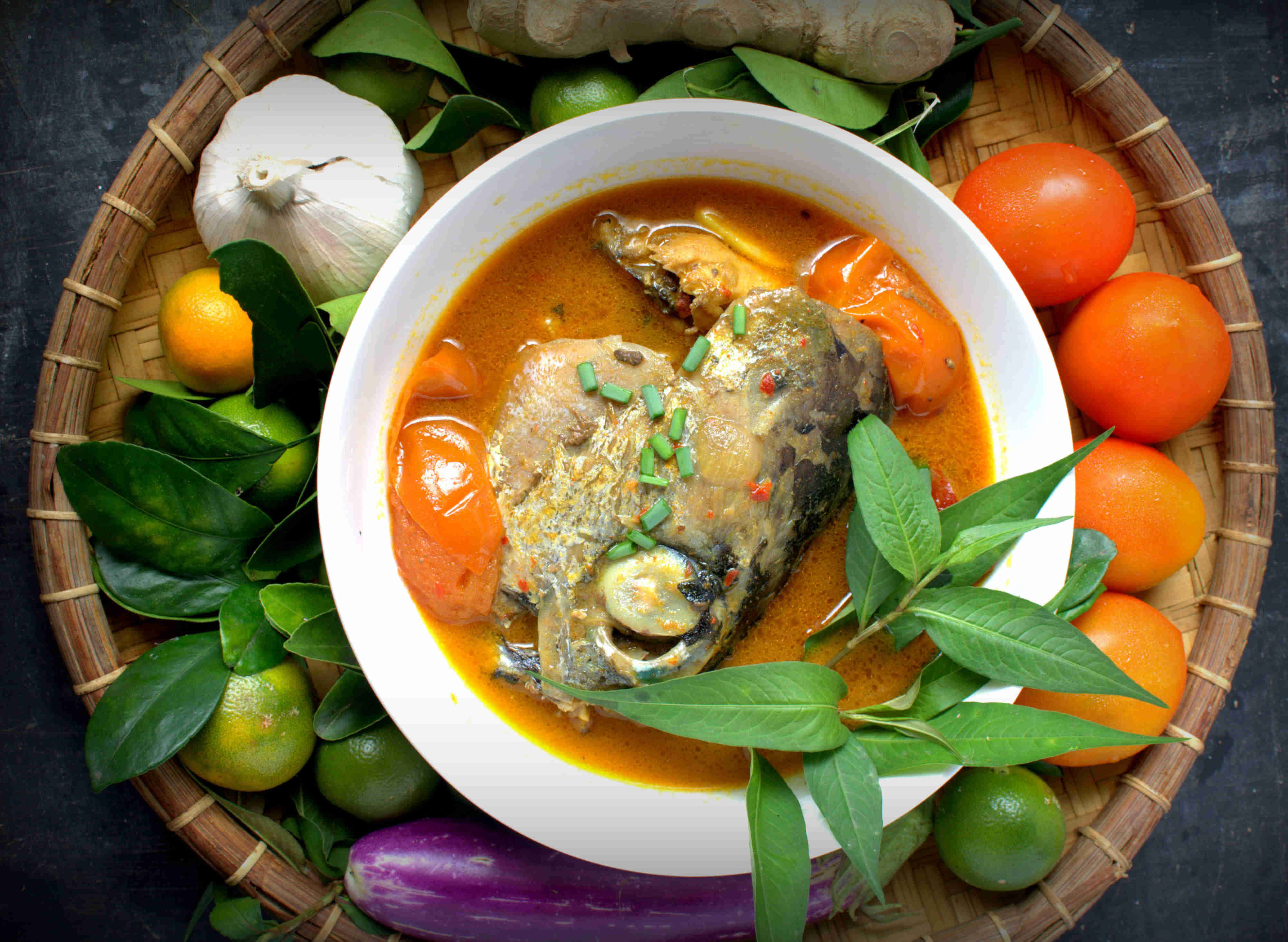 Food Photography 桌布 壁紙 — 一碗冒煙的魚咖哩，配上綠色香草，周圍是新鮮番茄、萊姆和大蒜，放在編織竹盤上。