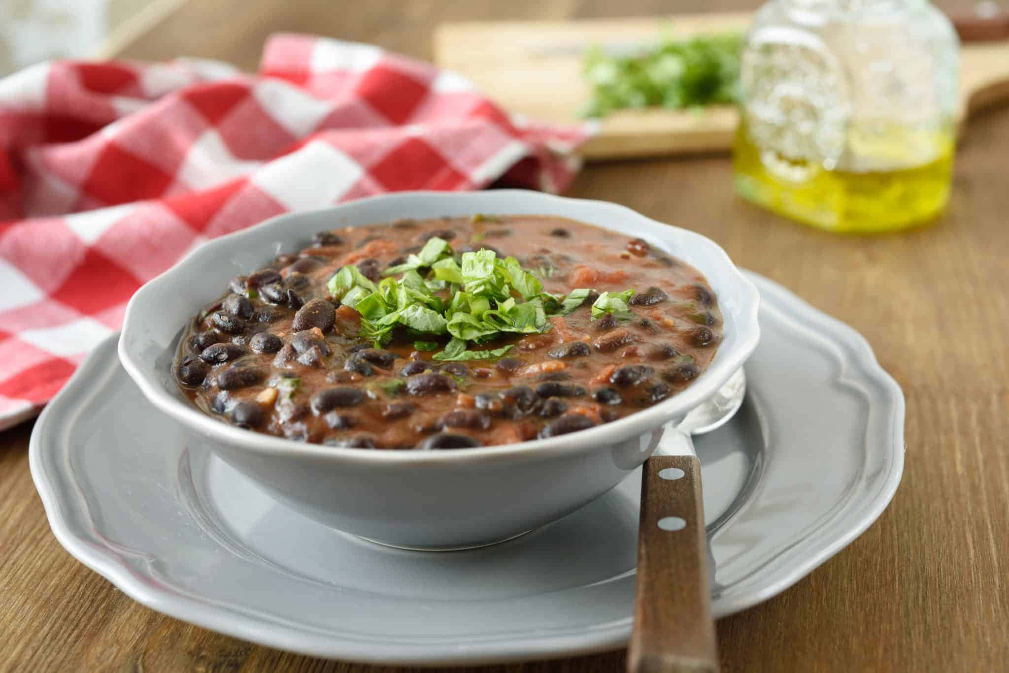 Black Bean Soup 桌布 壁紙 — 一碗冒煙的黑豆，上面放著新鮮香菜，放在灰色盤子上，旁邊有一張紅白格子餐巾。
