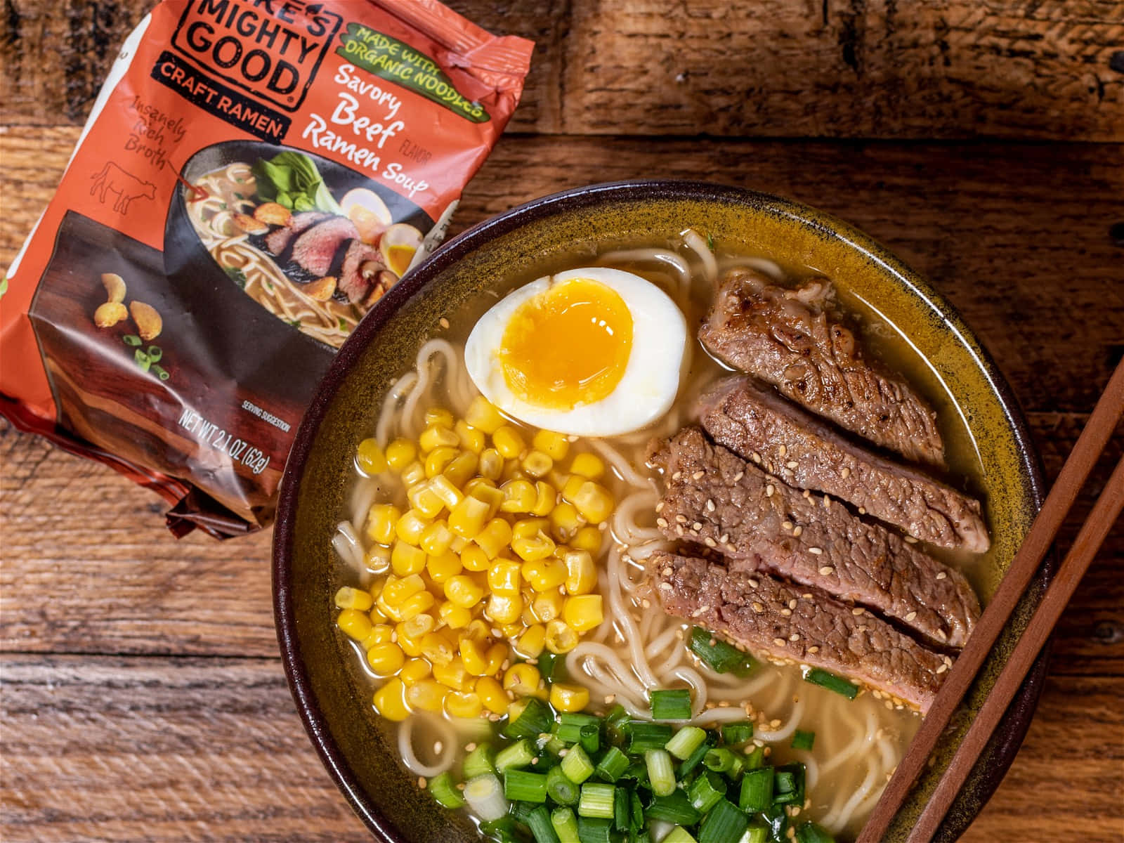 Noodles 桌布 壁紙 — 一碗冒著熱氣、滋味濃郁的牛肉拉麵，上面放著半熟蛋、玉米和青蔥，旁邊放著調味包，擱在質樸的木桌上。