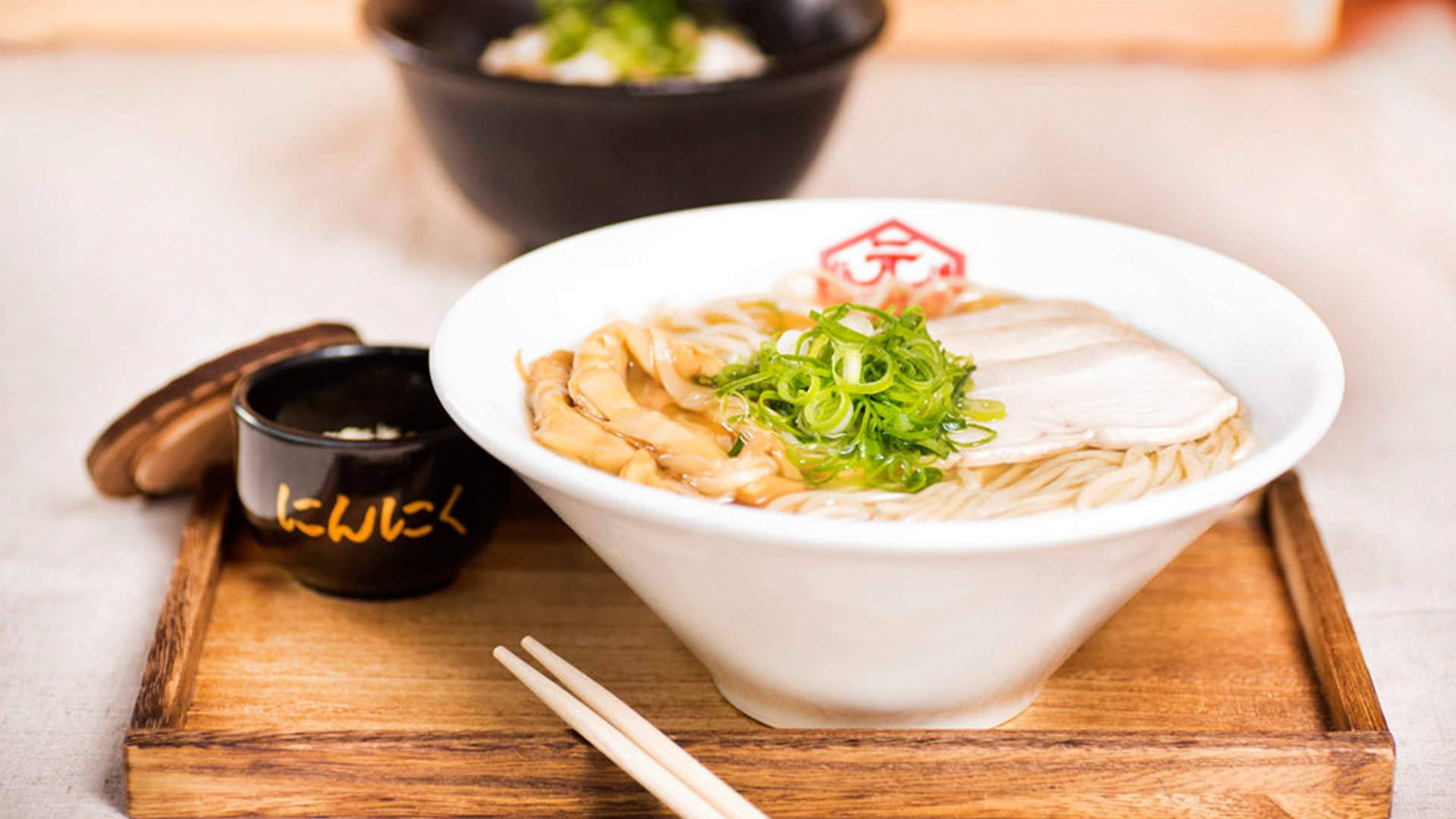 Ramen 桌布 壁紙 — 一碗冒著熱氣的拉麵，上面有蔥花和豬肉片，放在木托盤上，附有筷子和一個黑色小醬油碟。