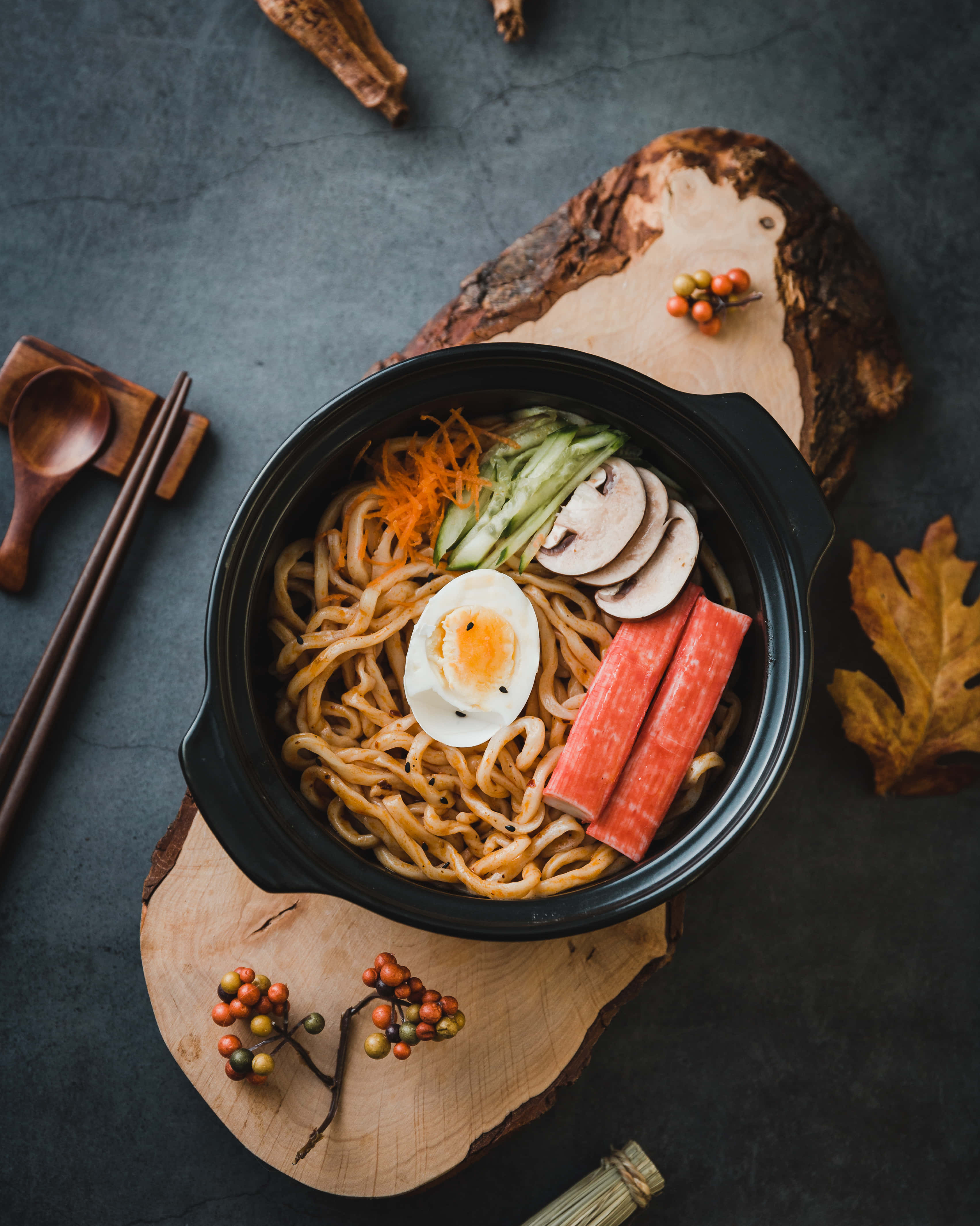 Noodles 桌布 壁紙 — 一碗冒著熱氣的拉麵，配有溏心蛋、切片蘑菇、胡蘿蔔絲和蟹棒，放在一個質樸的木板上，周圍環繞著秋葉和木製餐具。