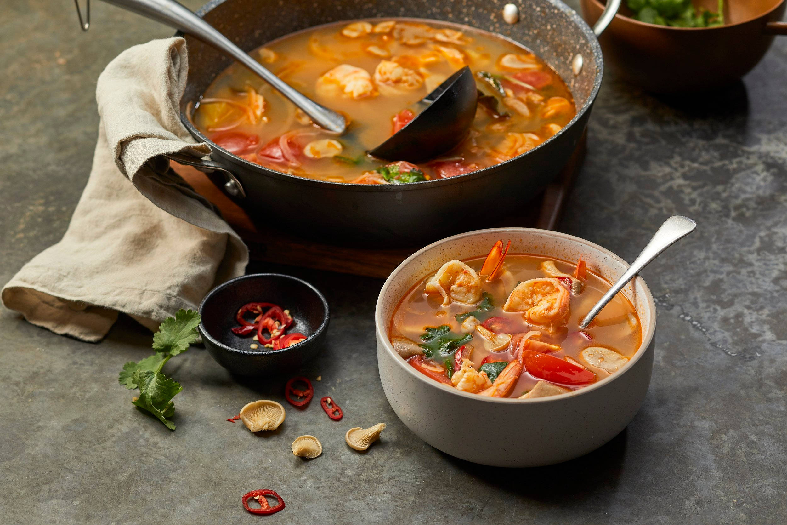 Delicious Food 桌布 壁紙 — 一碗冒著熱氣的泰式冬陰功湯，有蝦和辣椒，旁邊放著一個煎鍋，用新鮮香草和散落的紅辣椒裝飾在深色石面上。
