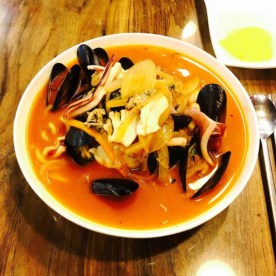 Food Photography 桌布 壁紙 — 一碗冒著熱氣的海鮮燉菜，有淡菜、魷魚和洋蔥，放在濃郁的橙色湯汁中，置於木桌上。