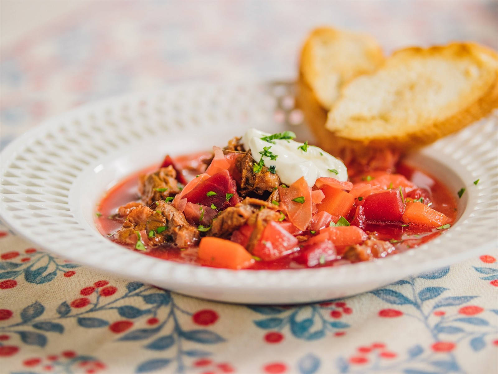 tomato 桌布 壁紙 — 一碗冒著熱氣的濃郁紅羅宋湯，上面放有酸奶油和新鮮香草，旁邊是金黃色的吐司，放在花卉圖案的桌布上。