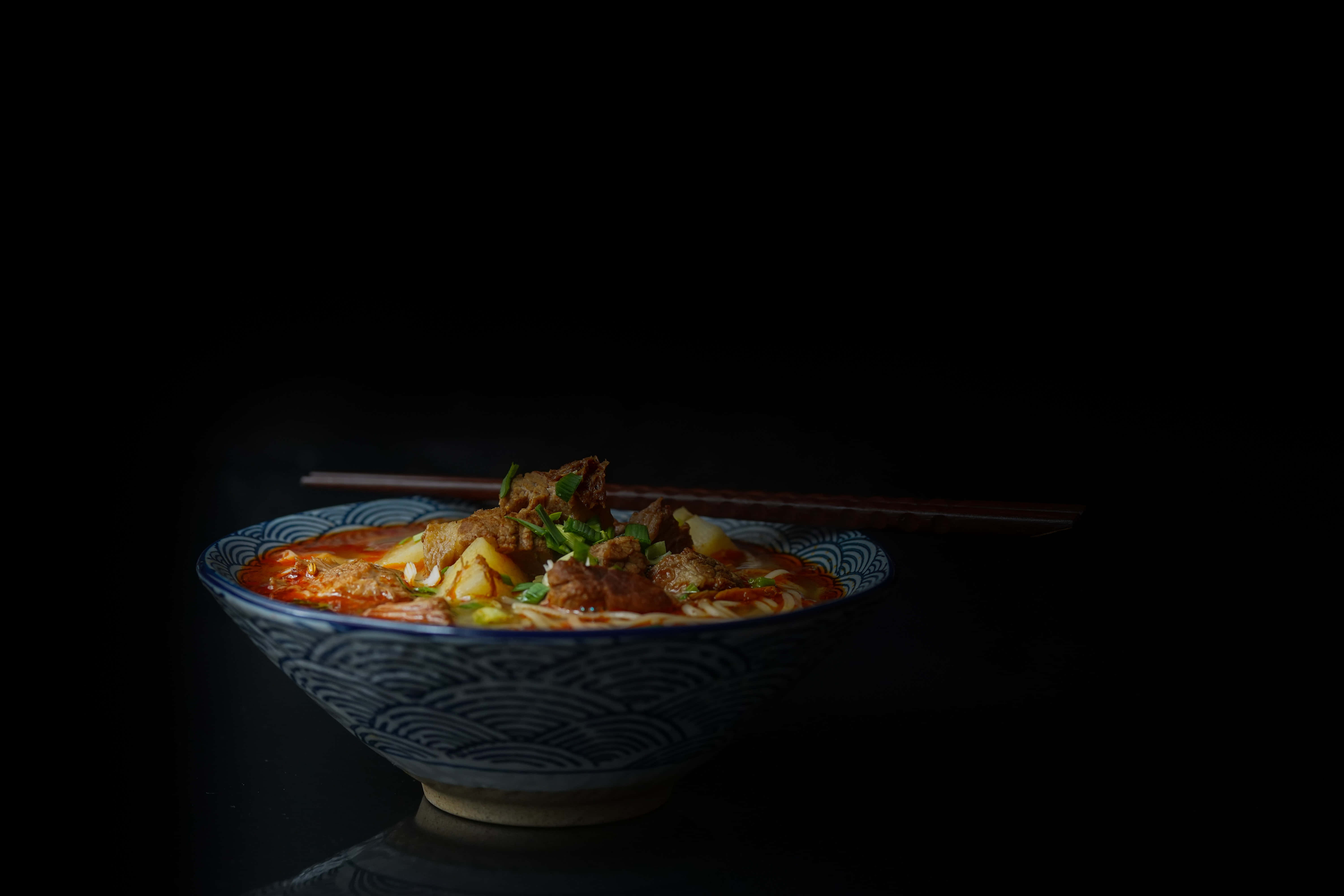 食物動圖 桌布 壁紙 — 一碗冒著熱氣的濃郁紅色湯麵，配上嫩肉和青蔥，在戲劇性的聚光燈下，在漆黑的背景中閃耀。