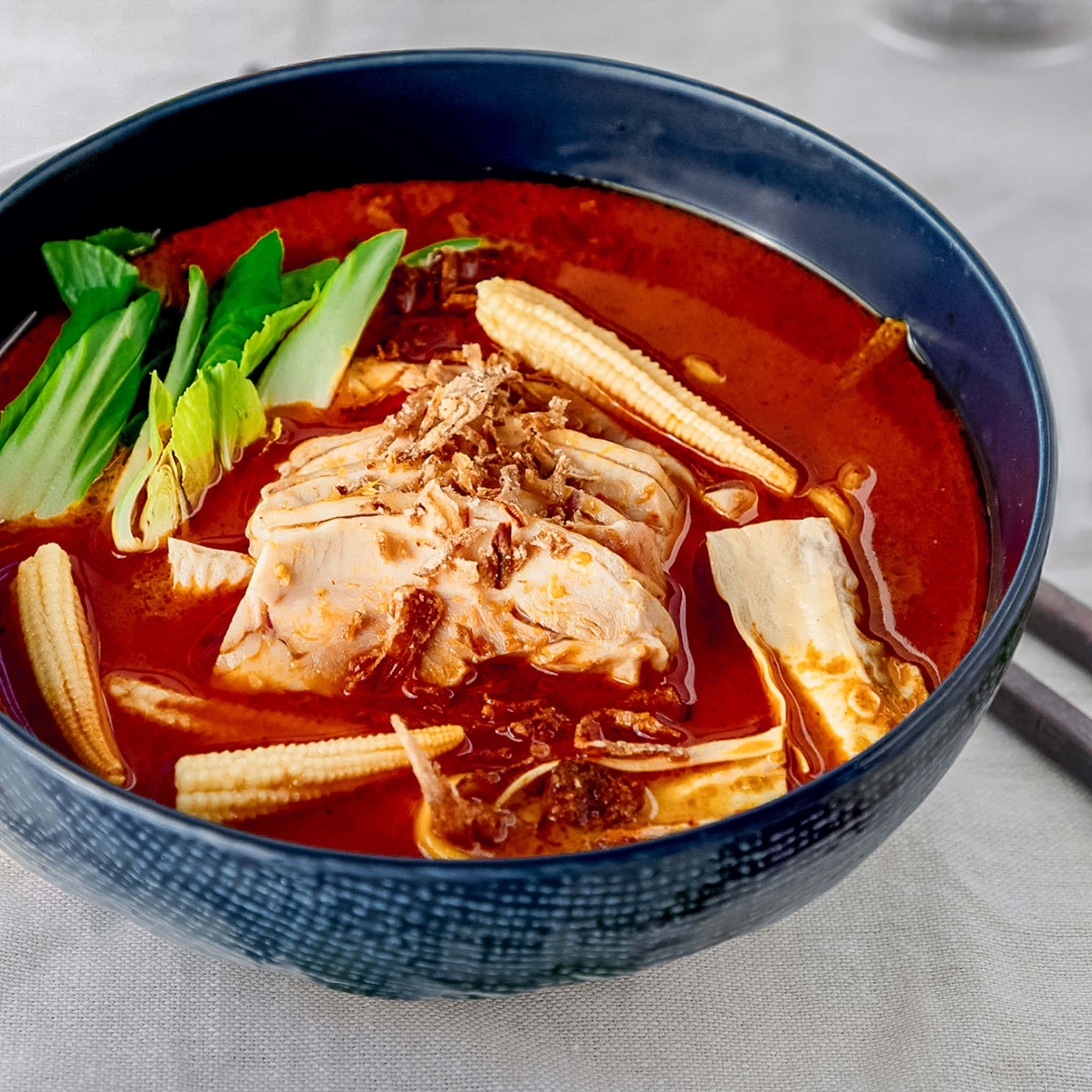Spicy 桌布 壁紙 — 一碗冒著熱氣的濃郁紅色辣湯，內有嫩雞肉、玉米筍和青江菜，盛載於質感豐富的深藍色陶瓷碗中。