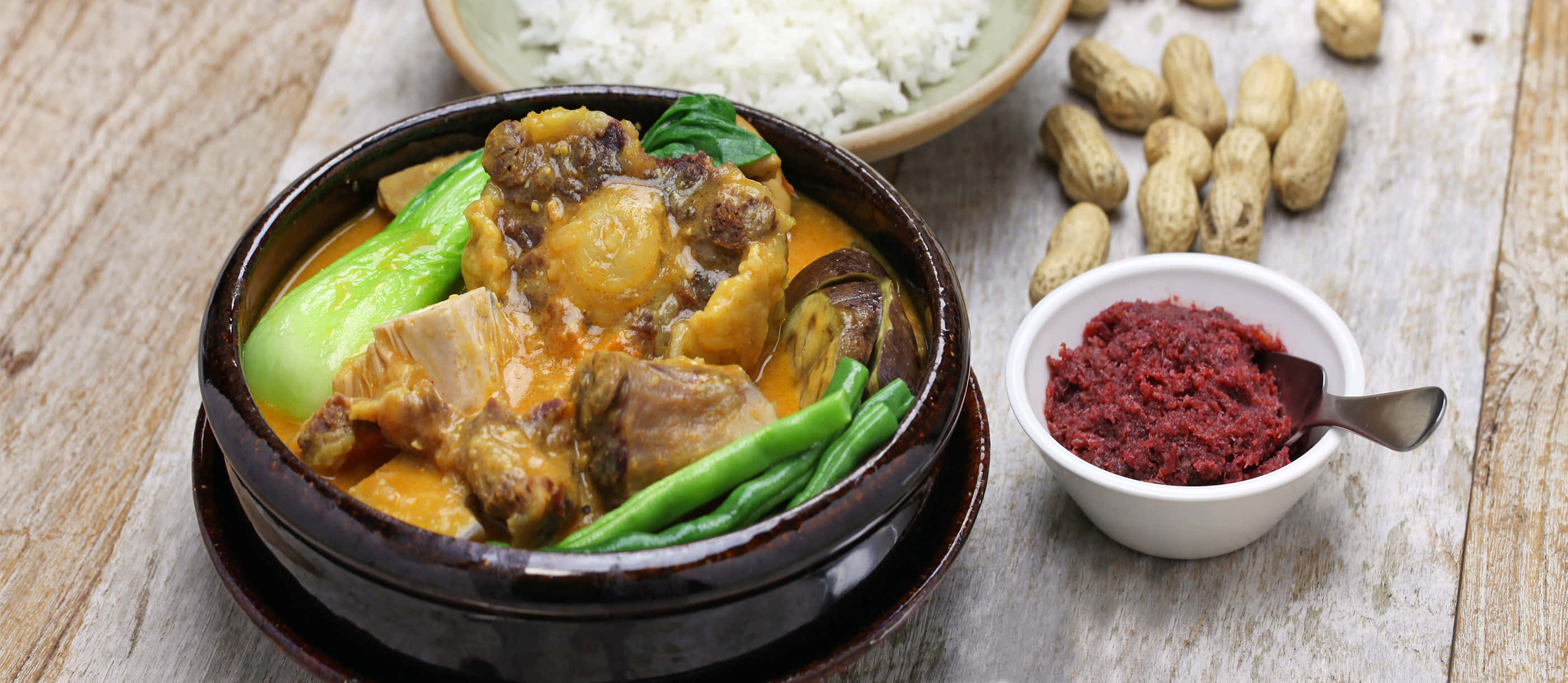 meat 桌布 壁紙 — 一碗冒著熱氣的濃郁黃咖哩，配有肉類和綠色蔬菜，旁邊放著白飯、花生和紅辣椒醬，擺在質樸的木桌上。