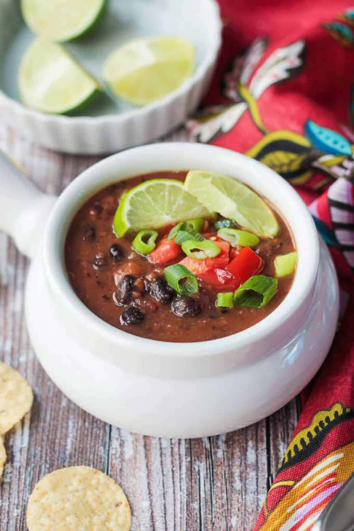 Black Bean Soup 桌布 壁紙 — 一碗冒著熱氣的濃郁黑豆湯，上面放著萊姆角、切碎的番茄和青蔥，放在一張質樸的木桌上，旁邊還有玉米片和一塊色彩繽紛的圖案布。