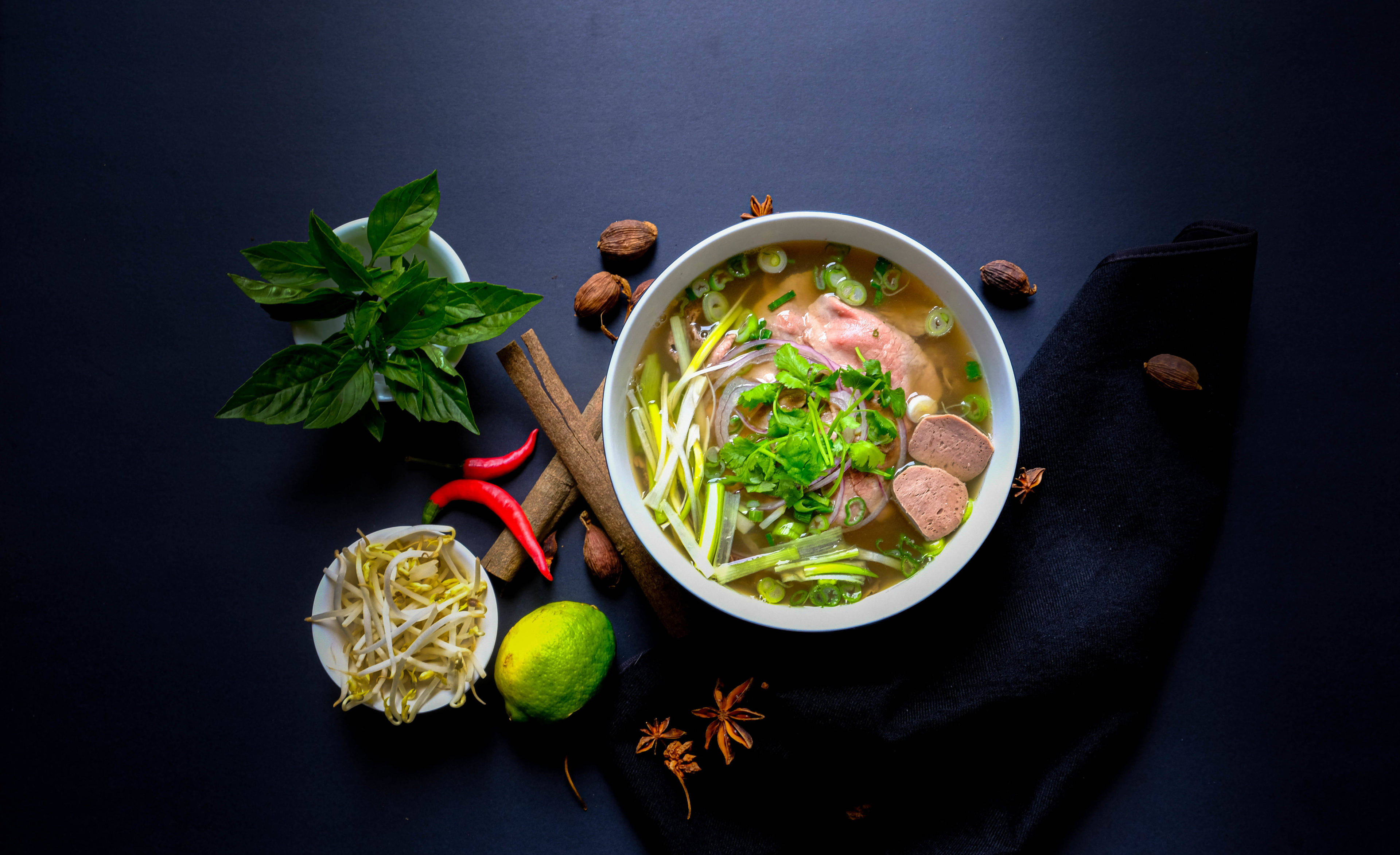 Food Photography 桌布 壁紙 — 一碗冒著熱氣的越南河粉，點綴著香草和肉，周圍是深色表面上的新鮮食材。