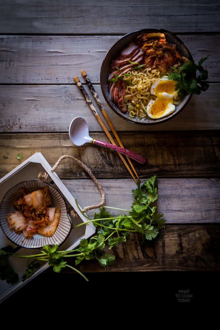 Noodles 桌布 壁紙 — 一碗冒著熱氣的韓國拉麵，搭配泡菜和半熟蛋，旁邊放著小菜和新鮮香菜，置於質樸的木桌上。