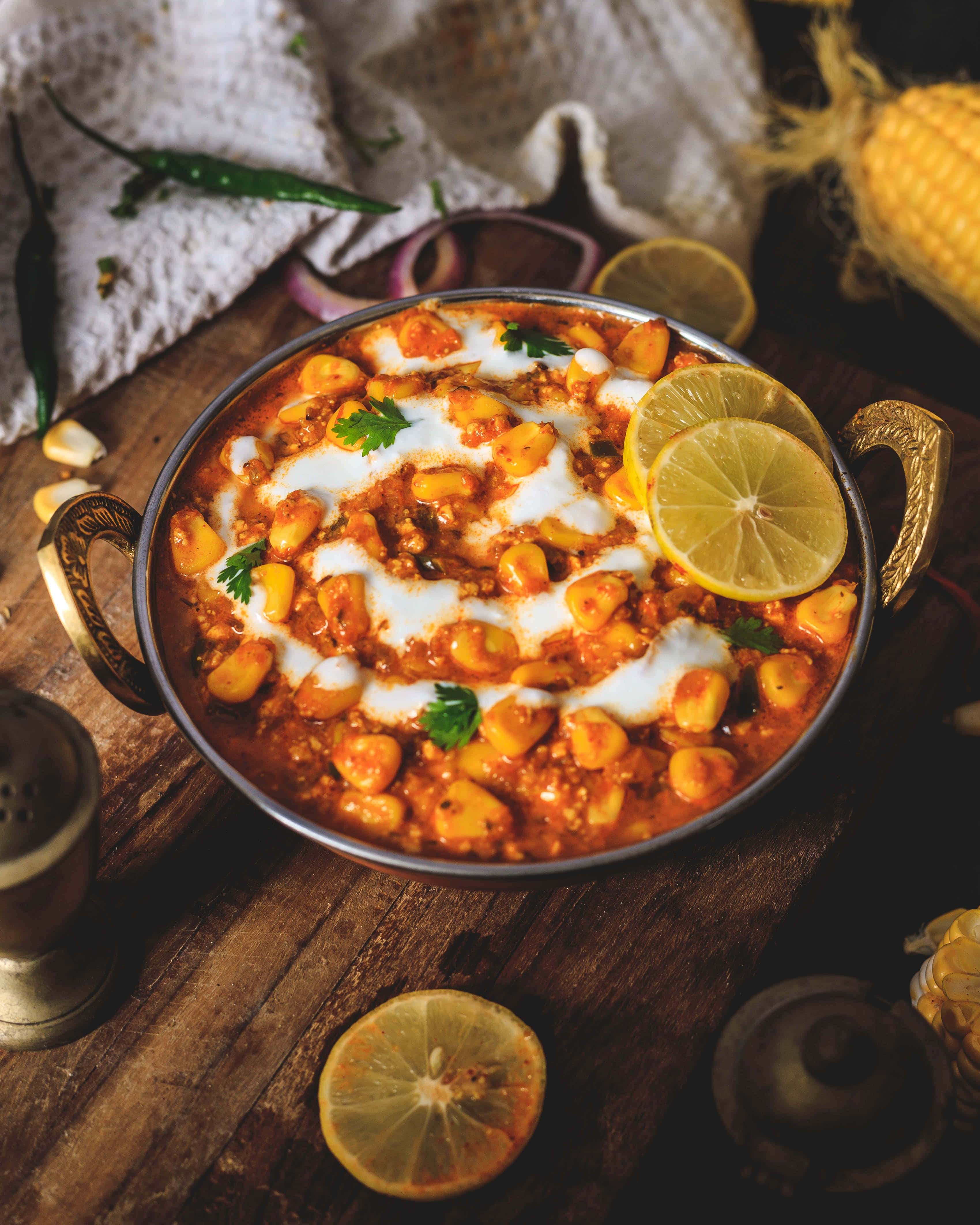 Indian 桌布 壁紙 — 一碗冒著熱氣的鮮橙色chole masala，上面點綴著優格、檸檬片和香菜，放在一個鄉村風格的木桌上，周圍環繞著香料和玉米。