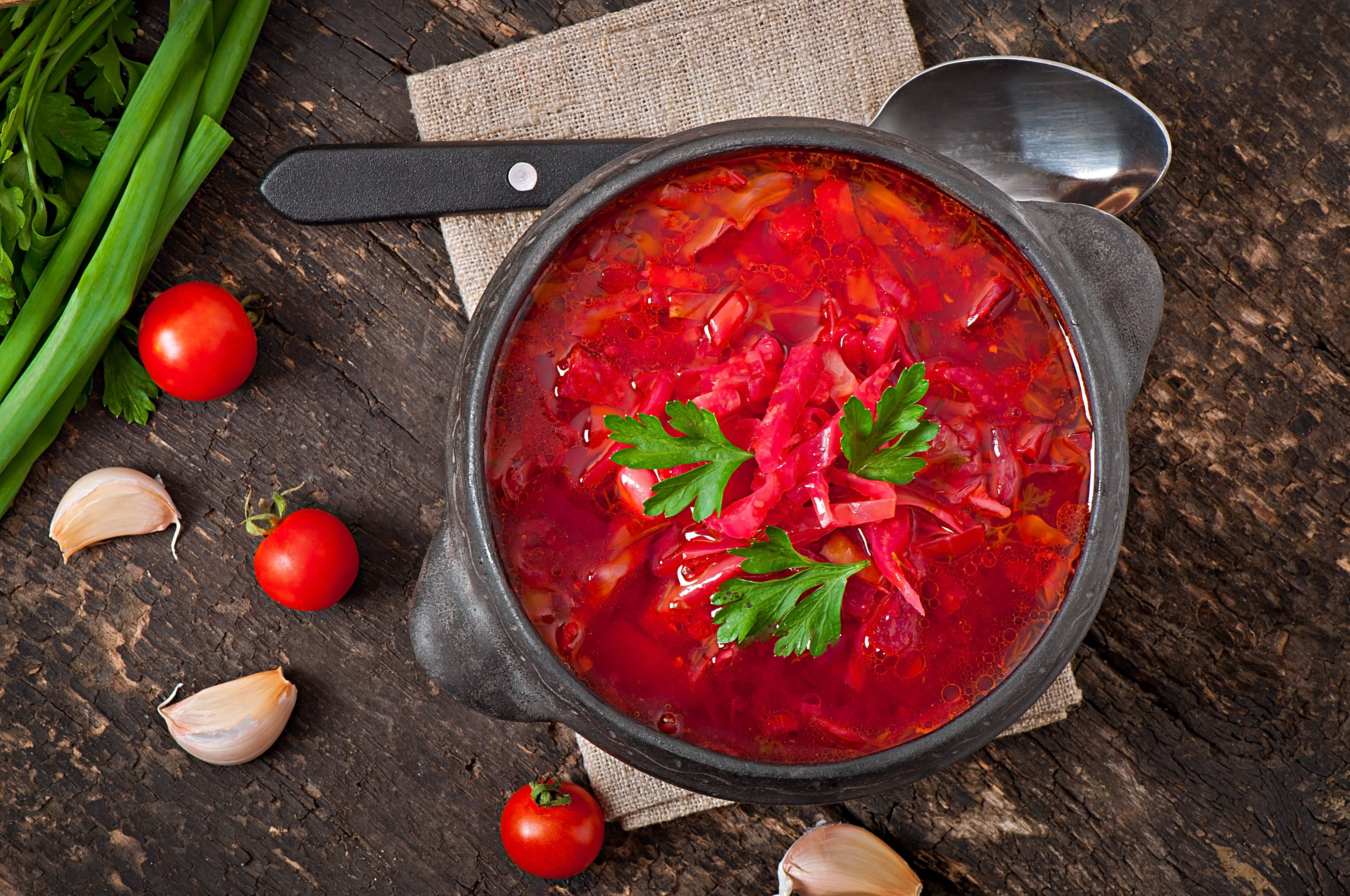 beetroot 桌布 壁紙 — 一碗冒著熱氣的鮮紅色羅宋湯，飾以歐芹，放在一張質樸的木桌上，周圍是新鮮的番茄、大蒜和青蔥。