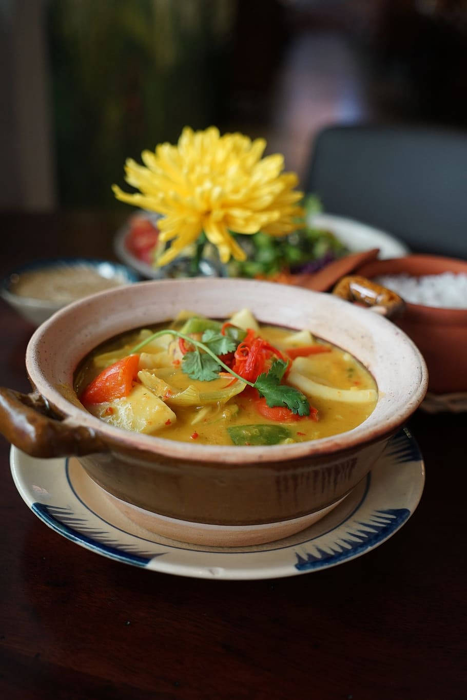 yummy 桌布 壁紙 — 一碗冒著熱氣的鮮黃色咖哩，配上新鮮香草和蔬菜，放在有圖案的盤子上，旁邊有一朵鮮黃色的花。