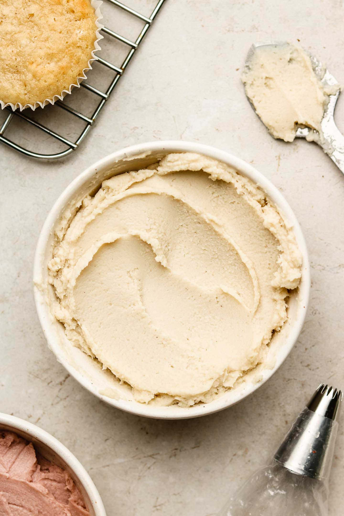 Buttercream 桌布 壁紙 — 一碗奶油米色糖霜放在淺色檯面上，旁邊有一個正在冷卻的杯子蛋糕和擠花工具。
