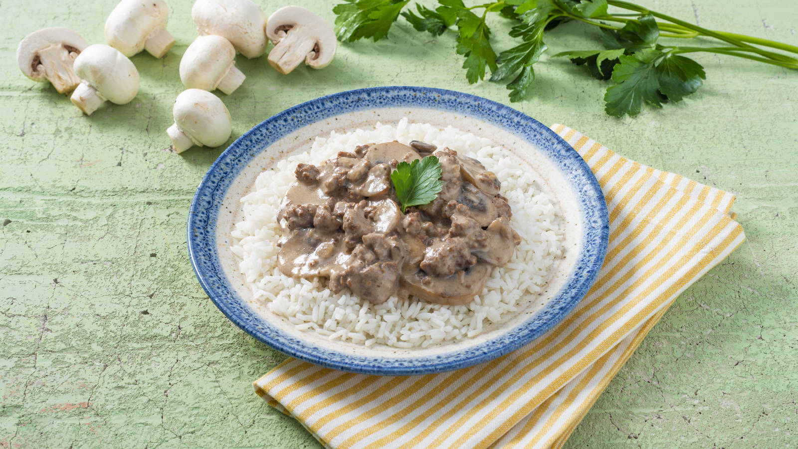mushrooms 桌布 壁紙 — 一碗奶油蘑菇牛肉燉飯放在白飯上，飾以歐芹，置於條紋餐巾上，背景為有紋理的綠色。