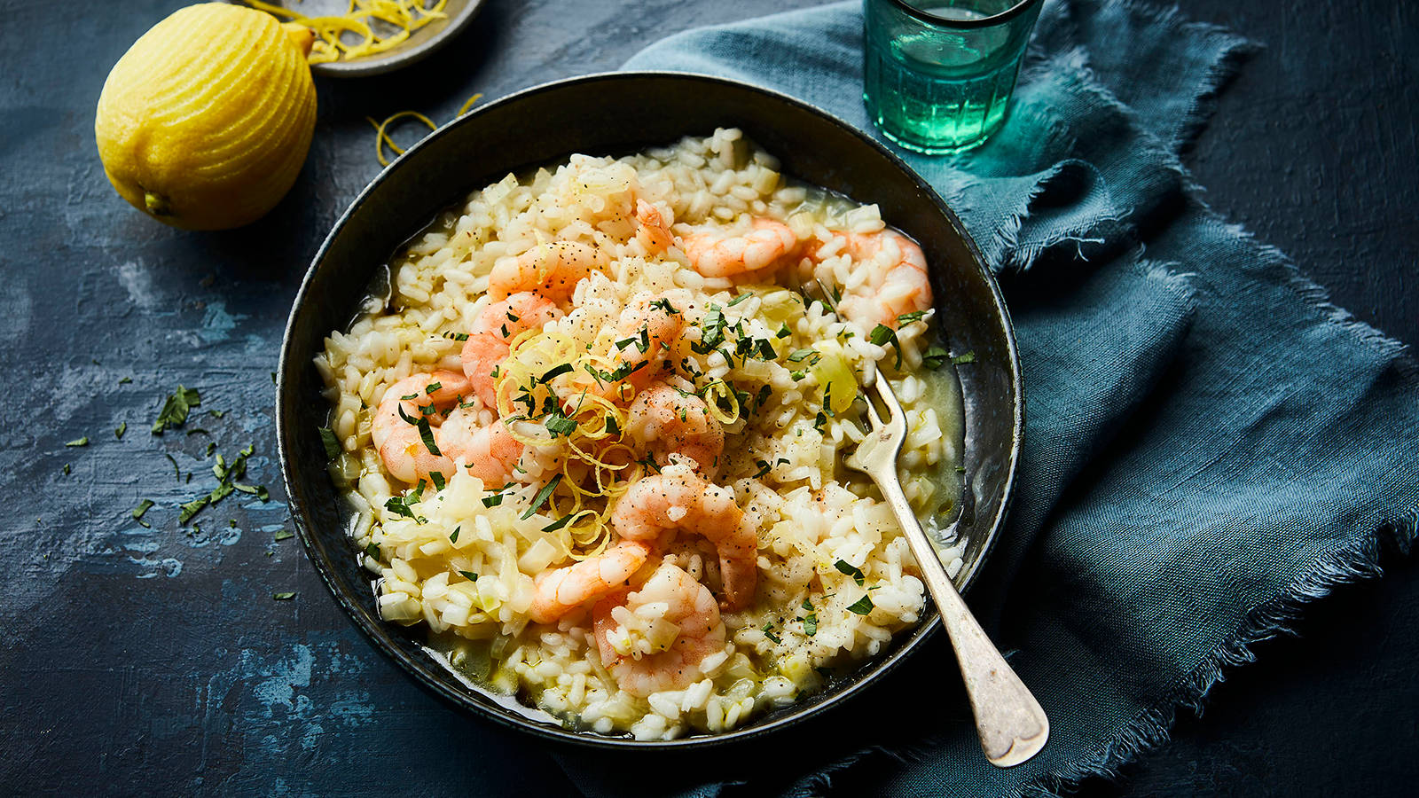 prawn 桌布 壁紙 — 一碗奶油蝦肉燉飯，飾以檸檬皮和香草，放在深藍色布上，旁邊有整個檸檬和綠色玻璃杯。