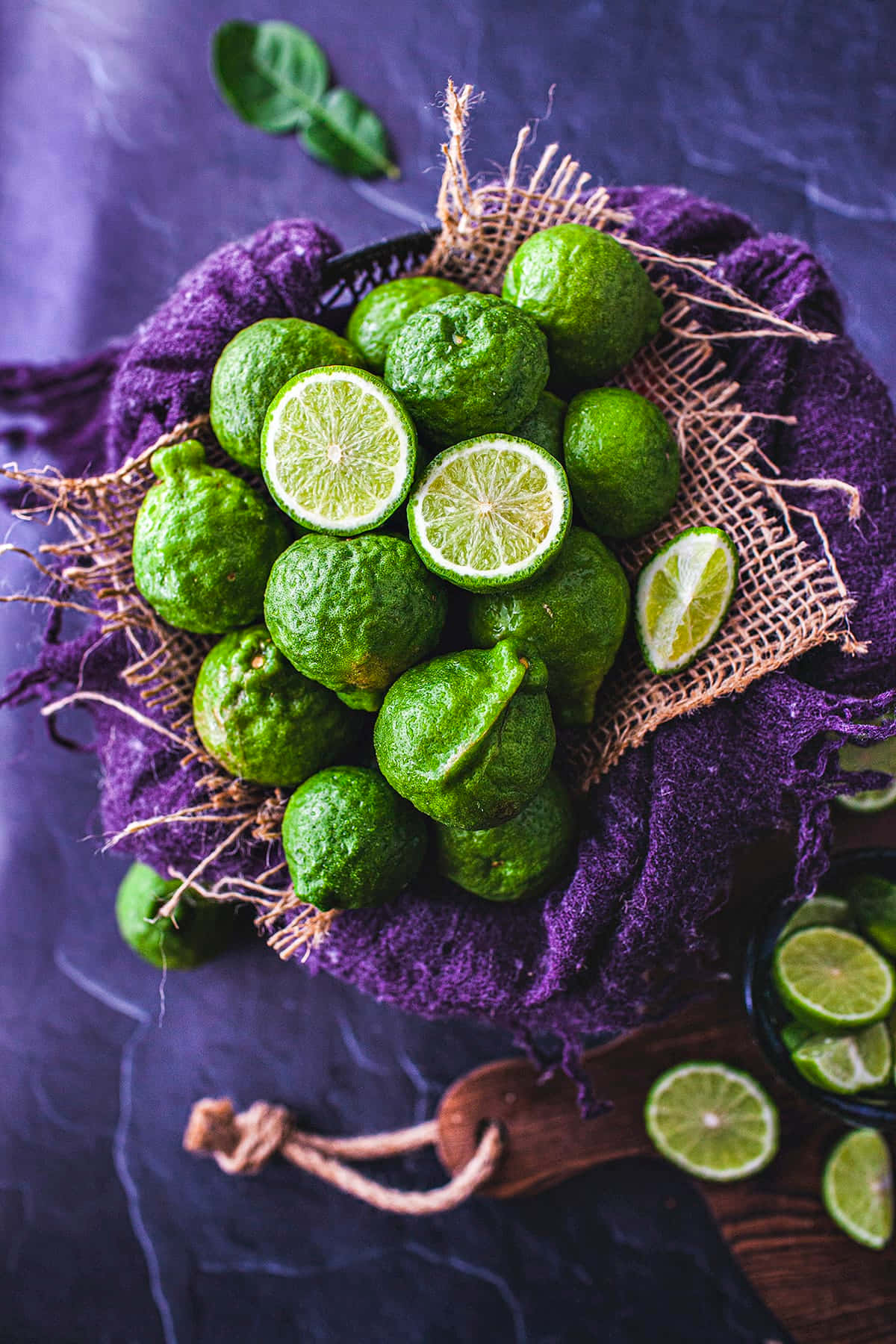 citrus 桌布 壁紙 — 一碗新鮮青檸放在紫色布料和麻布上，襯托深色紋理背景，色彩鮮明。