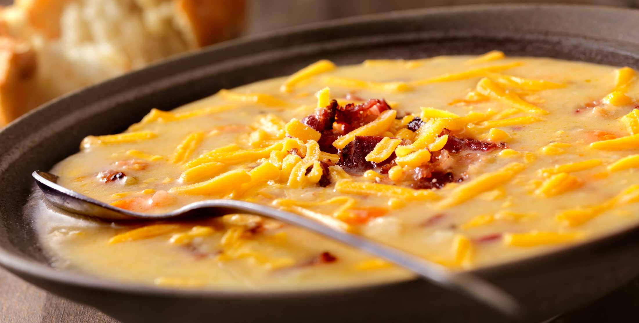 Chowder 桌布 壁紙 — 一碗深色碗中的奶油起司漢堡湯，上面撒有切達起司絲和脆培根，配有一隻湯匙。