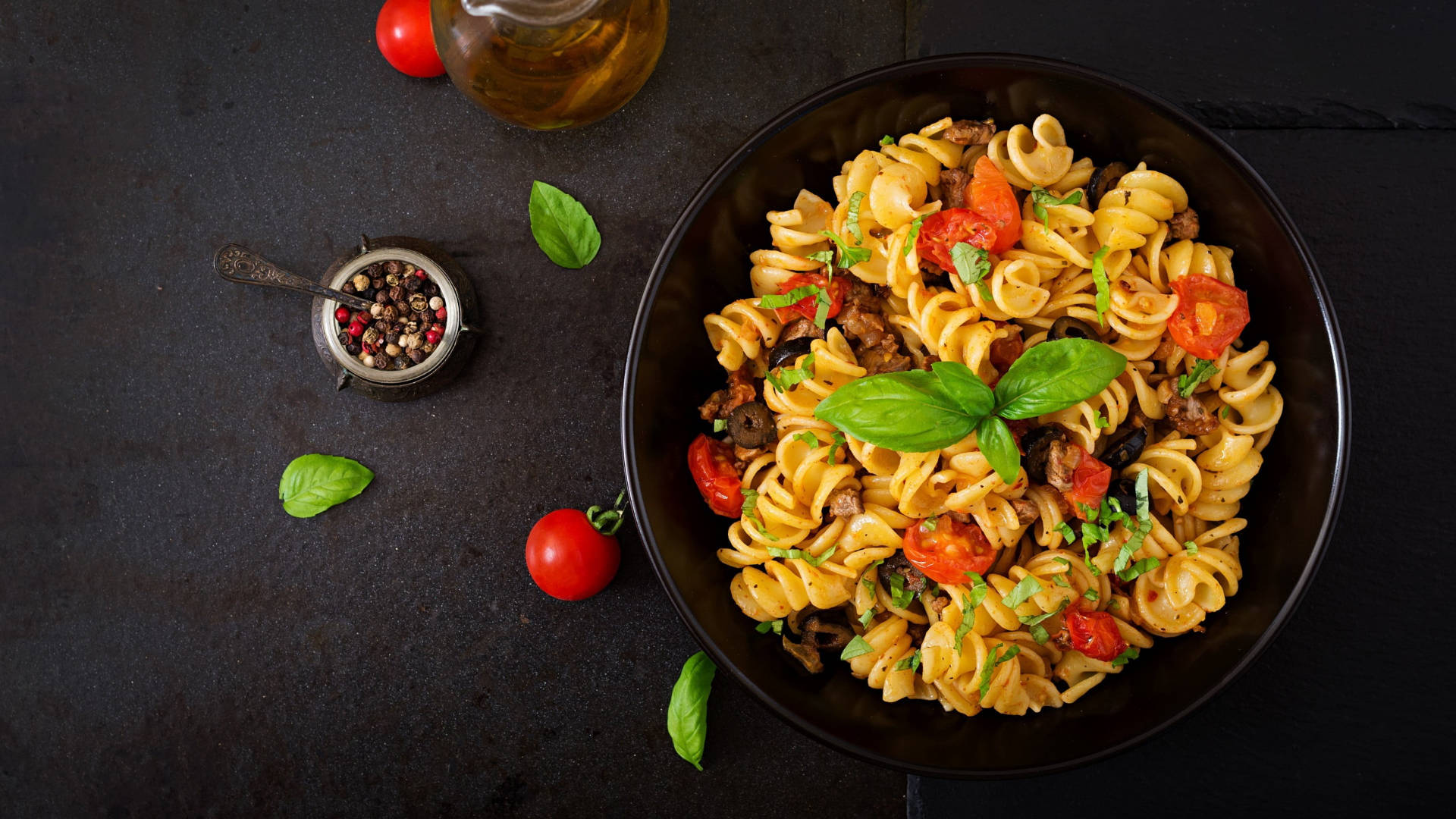 Pasta 桌布 壁紙 — 一碗深色螺旋義大利麵搭配番茄、羅勒和肉，放在質樸的黑色表面上，周圍環繞著新鮮食材。