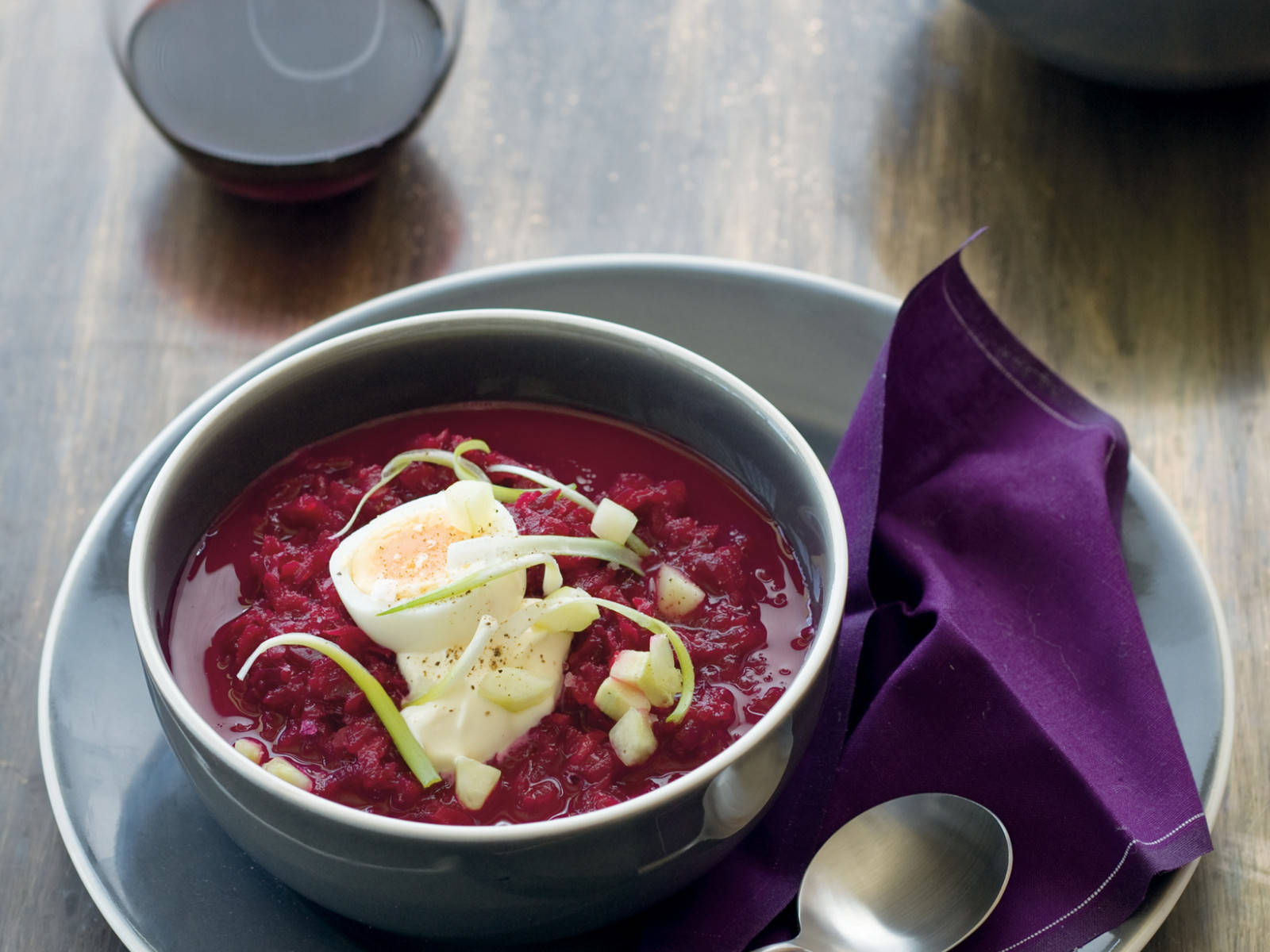 beetroot 桌布 壁紙 — 一碗濃郁的深紅色甜菜湯，飾以半個水煮蛋和切碎的蘋果，放在灰色碗中，置於木桌上，旁邊有紫色餐巾和湯匙。
