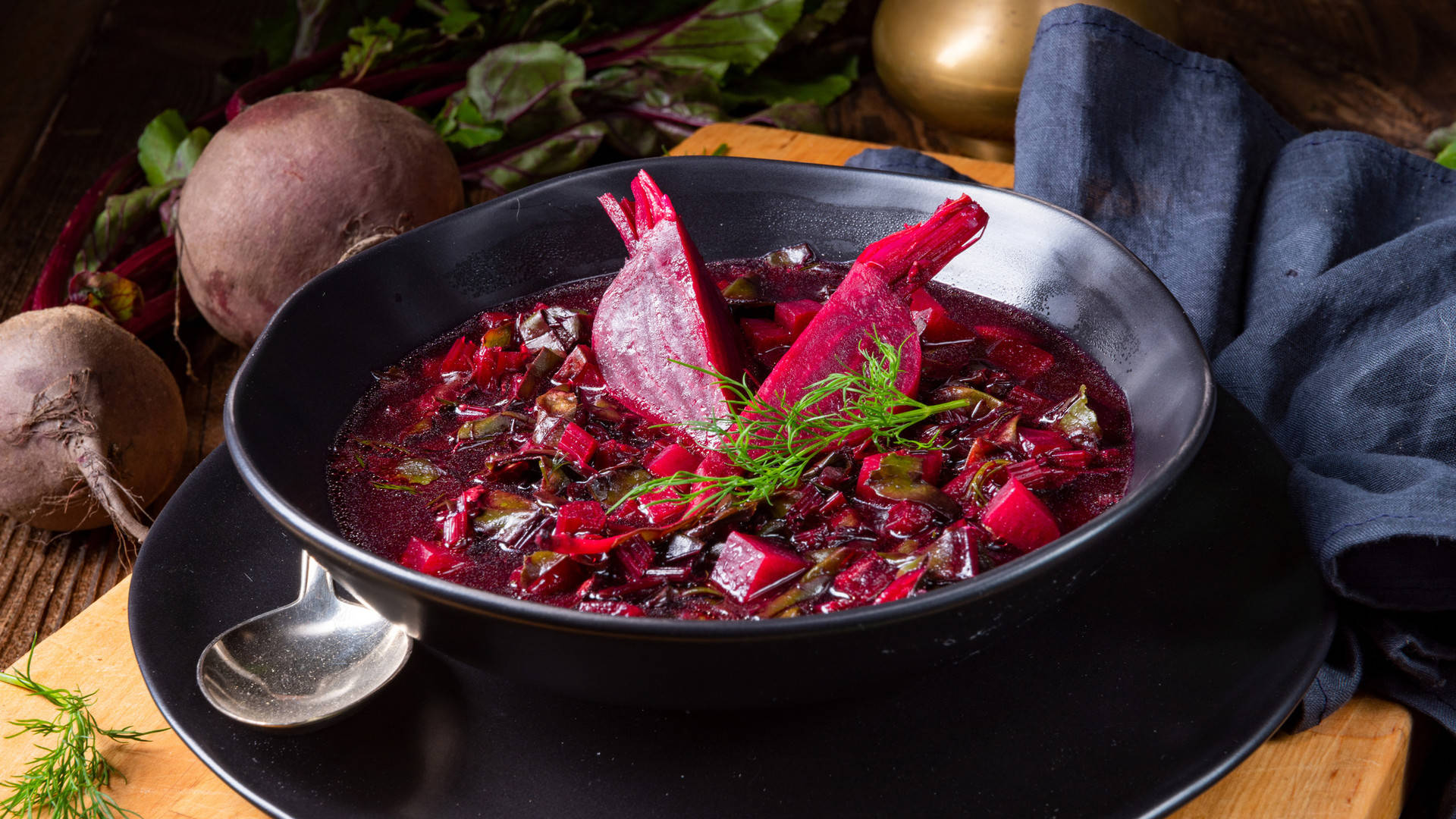 beetroot 桌布 壁紙 — 一碗濃郁的深紅色羅宋湯，飾以甜菜片和蒔蘿，放在黑色碗中，旁邊有整個甜菜和藍色布，置於木桌上。