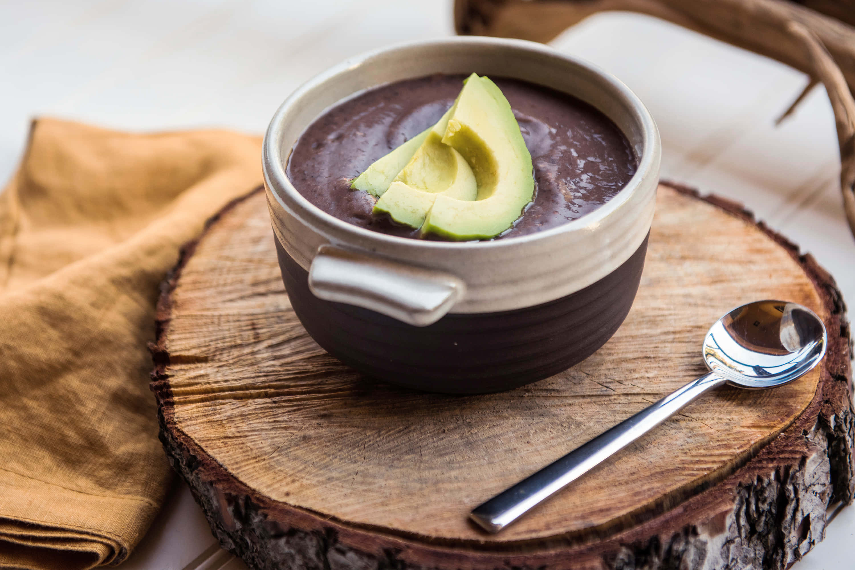 Black Bean Soup 桌布 壁紙 — 一碗濃郁的深色湯放在陶瓷碗中，上面鋪上新鮮酪梨片，放在一塊鄉村風格的木板上，旁邊放著湯匙。
