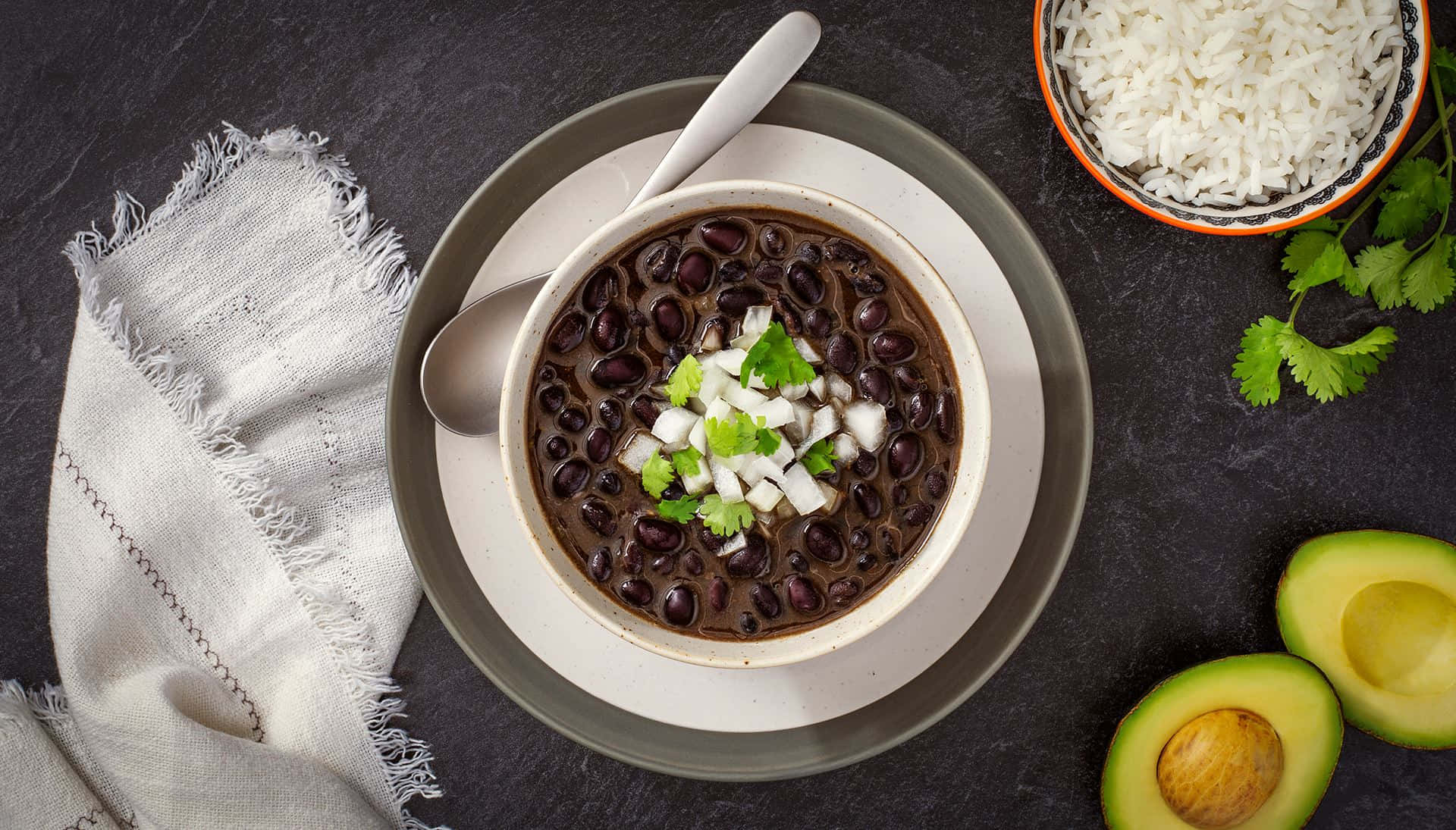 Black Bean Soup 桌布 壁紙 — 一碗濃郁的黑豆，上面放上新鮮香菜和切碎的洋蔥，旁邊放著白飯、酪梨和亞麻餐巾，置於深色石板表面上。