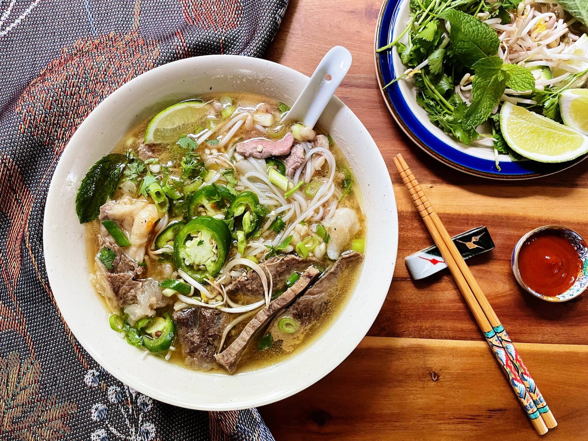 Noodles 桌布 壁紙 — 一碗熱氣騰騰的越南河粉，配上嫩牛肉河粉、萊姆片和新鮮香草，旁邊放著一碟青菜和色彩繽紛的筷子，置於木桌上。