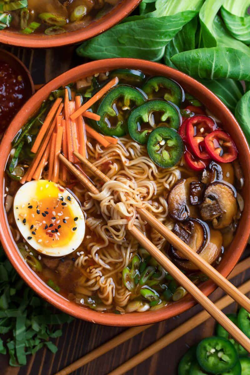 Noodles 桌布 壁紙 — 一碗熱騰騰的拉麵，上面放著半熟蛋、切片墨西哥辣椒、蘑菇和紅蘿蔔，周圍環繞著新鮮蔬菜。
