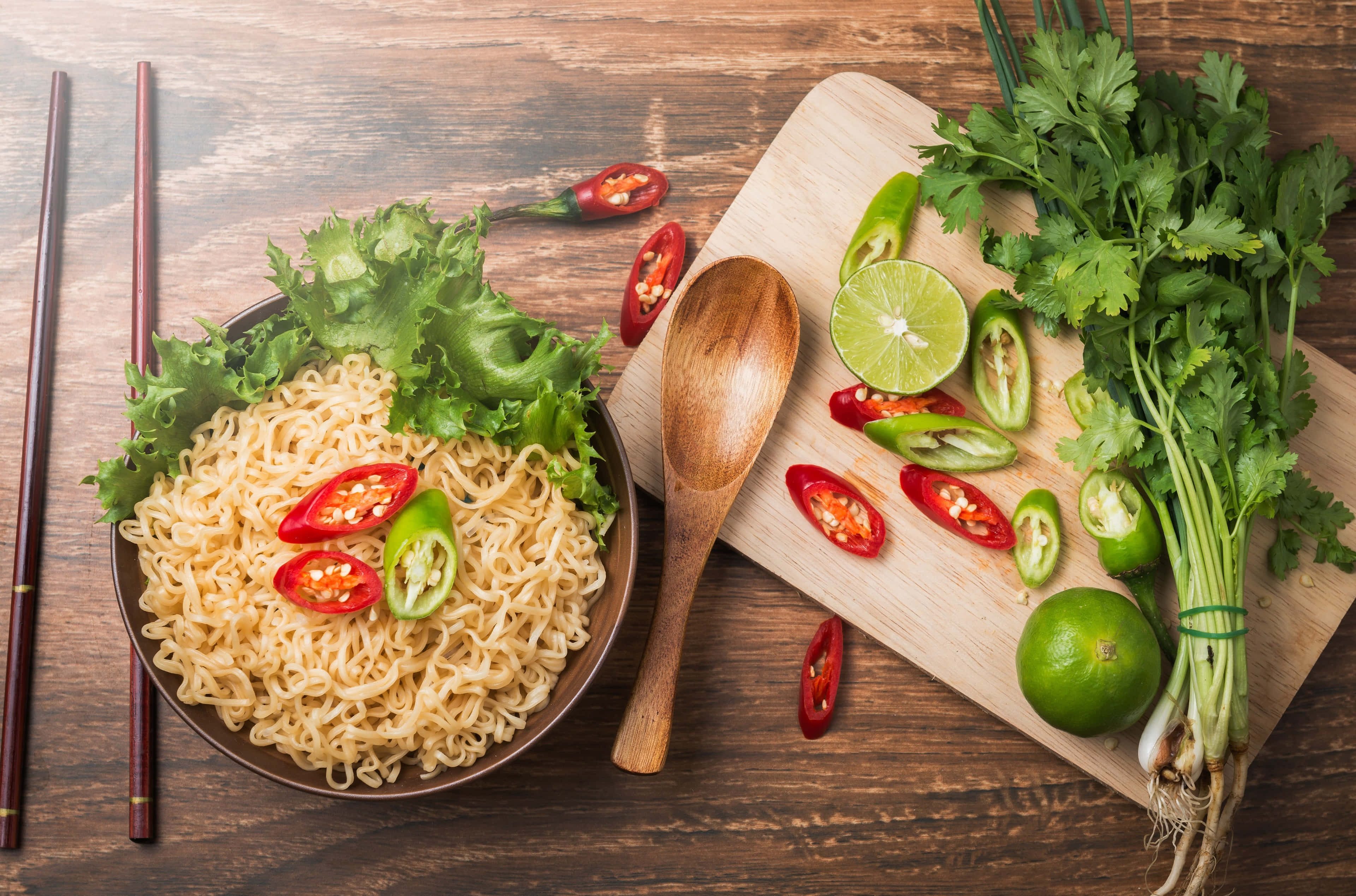 4K Pasta 桌布 壁紙 — 一碗熱騰騰的拉麵，上面鋪上新鮮生菜、切片紅綠辣椒，旁邊放著一個木製砧板，上面有萊姆、香菜和整條辣椒，放在一張質樸的木桌上。