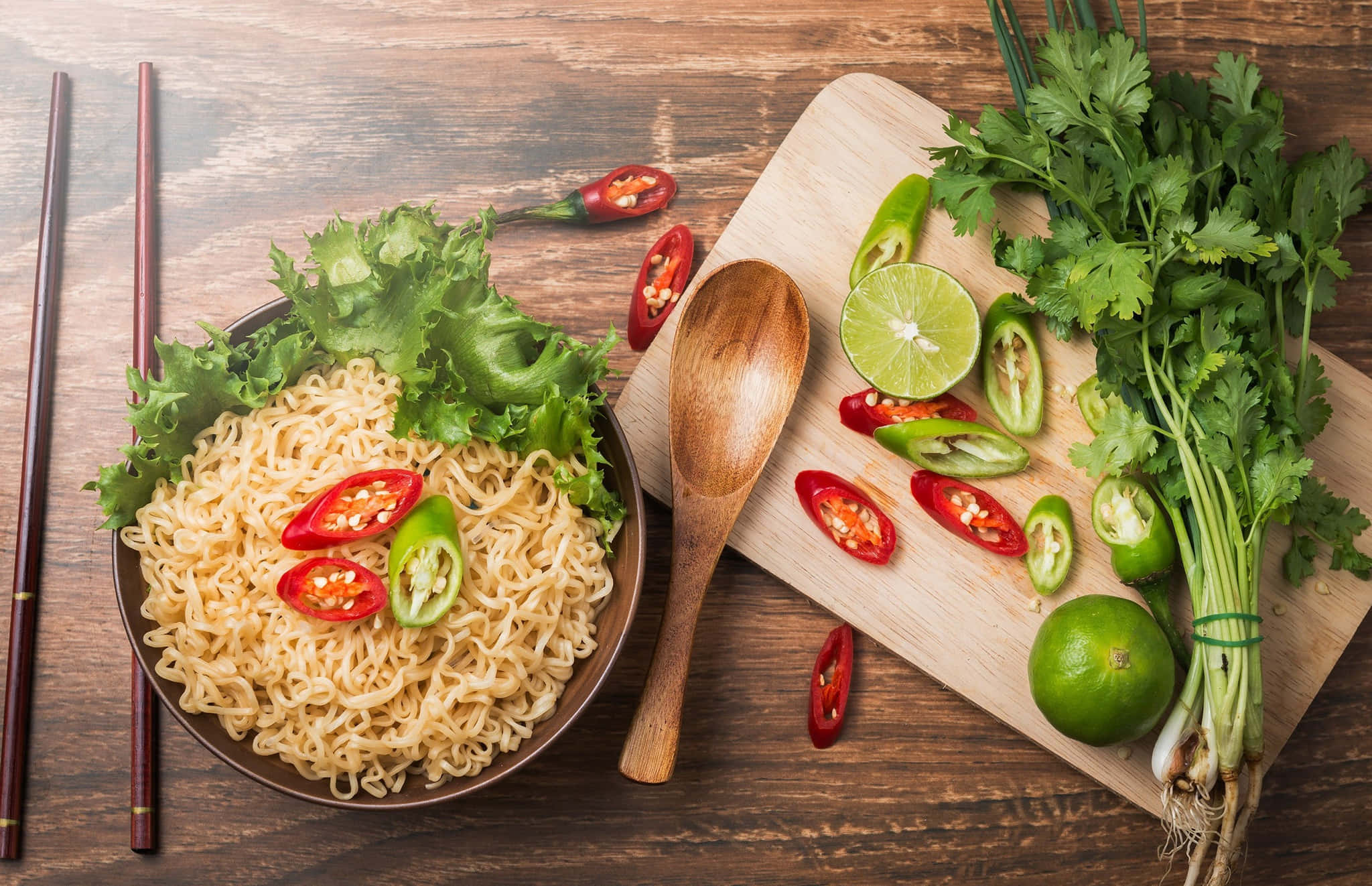 Food Photography 桌布 壁紙 — 一碗熱騰騰的拉麵，上面鋪上新鮮生菜和切片紅綠辣椒，旁邊放著一塊木砧板，上面有香菜、萊姆和更多辣椒，置於鄉村風木桌上。