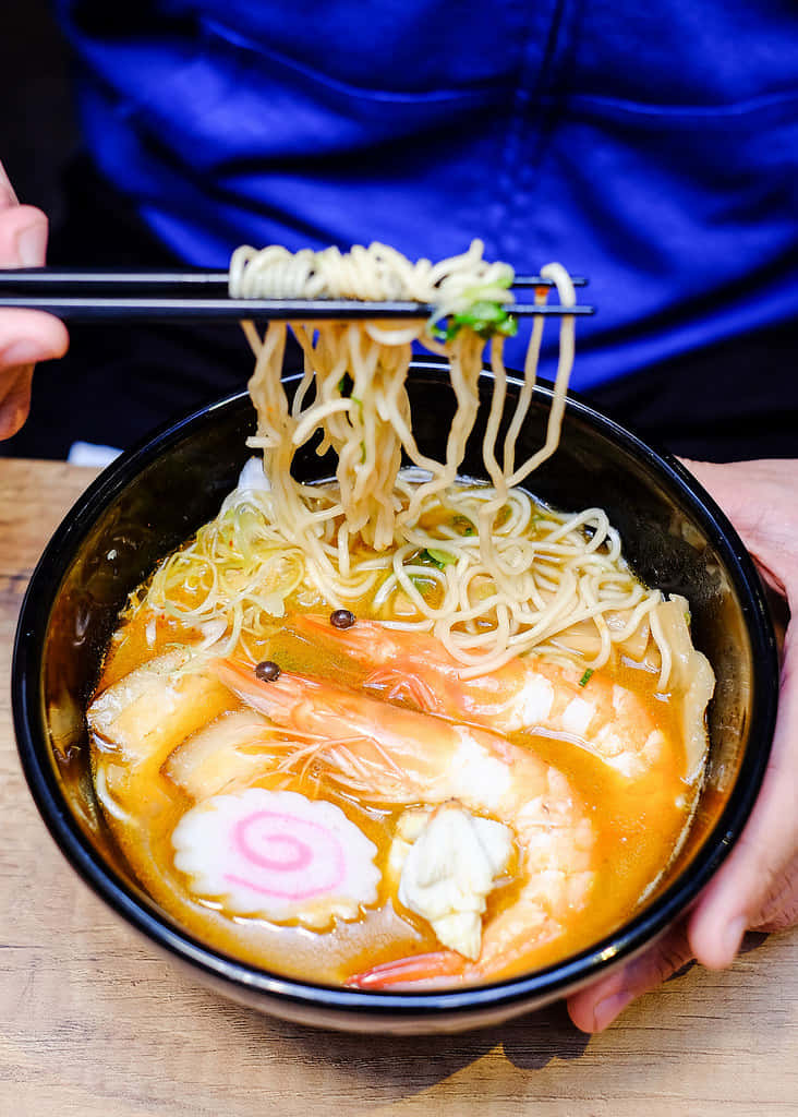 Noodles 桌布 壁紙 — 一碗熱騰騰的拉麵，有蝦和粉紅色漩渦魚板，正被筷子夾起。