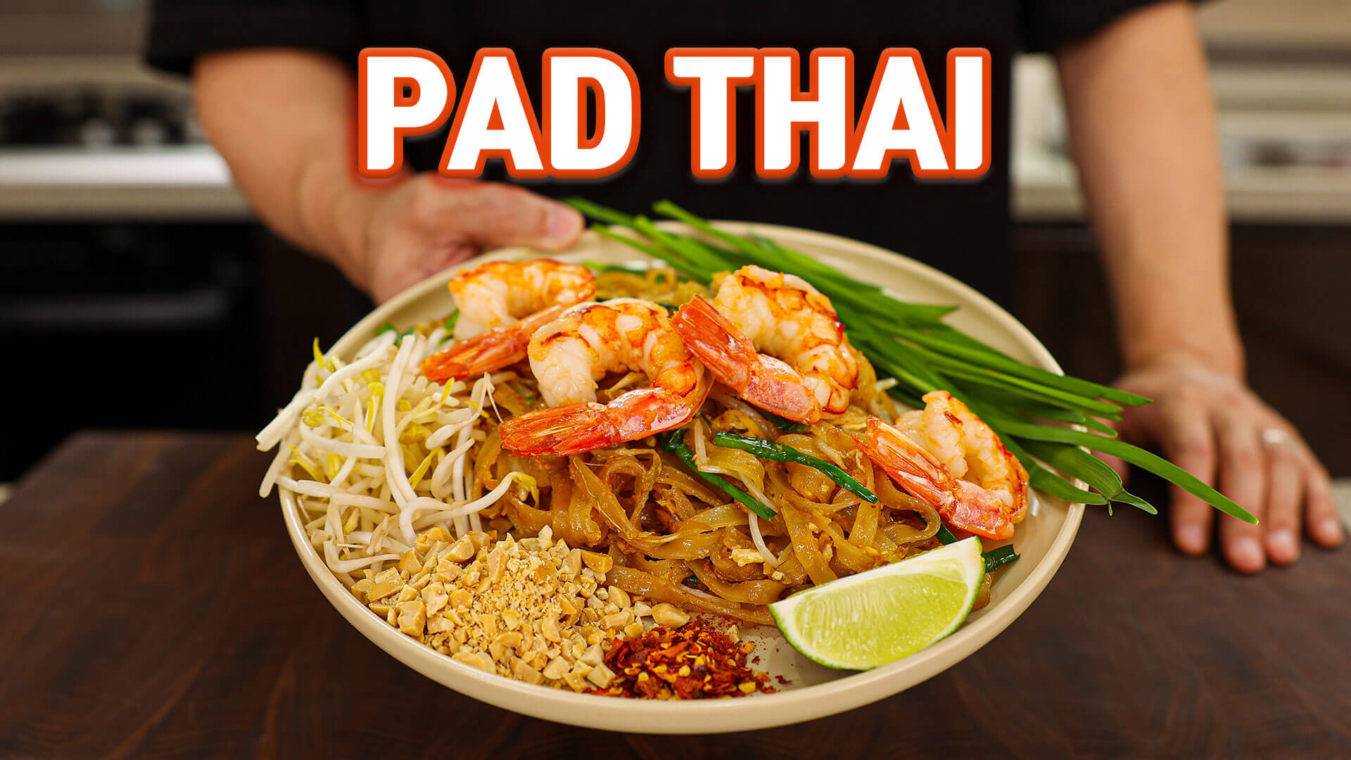 thai 桌布 壁紙 — 一碗熱騰騰的泰式炒河粉，上面放著蝦、豆芽、花生、辣椒片和青檸角，由廚師在廚房裡端著。