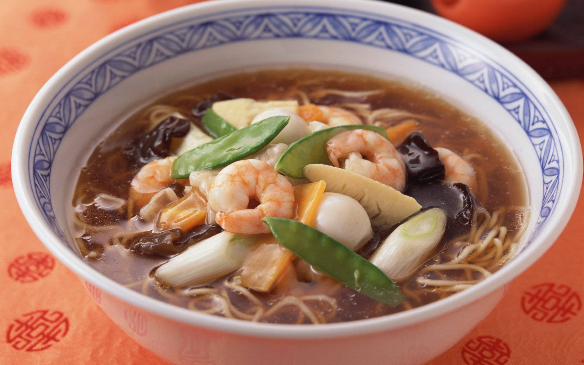 4K Food 桌布 壁紙 — 一碗熱騰騰的濃湯麵，配上蝦仁、荷蘭豆、蘑菇和豆腐，放在橙色圖案布上。
