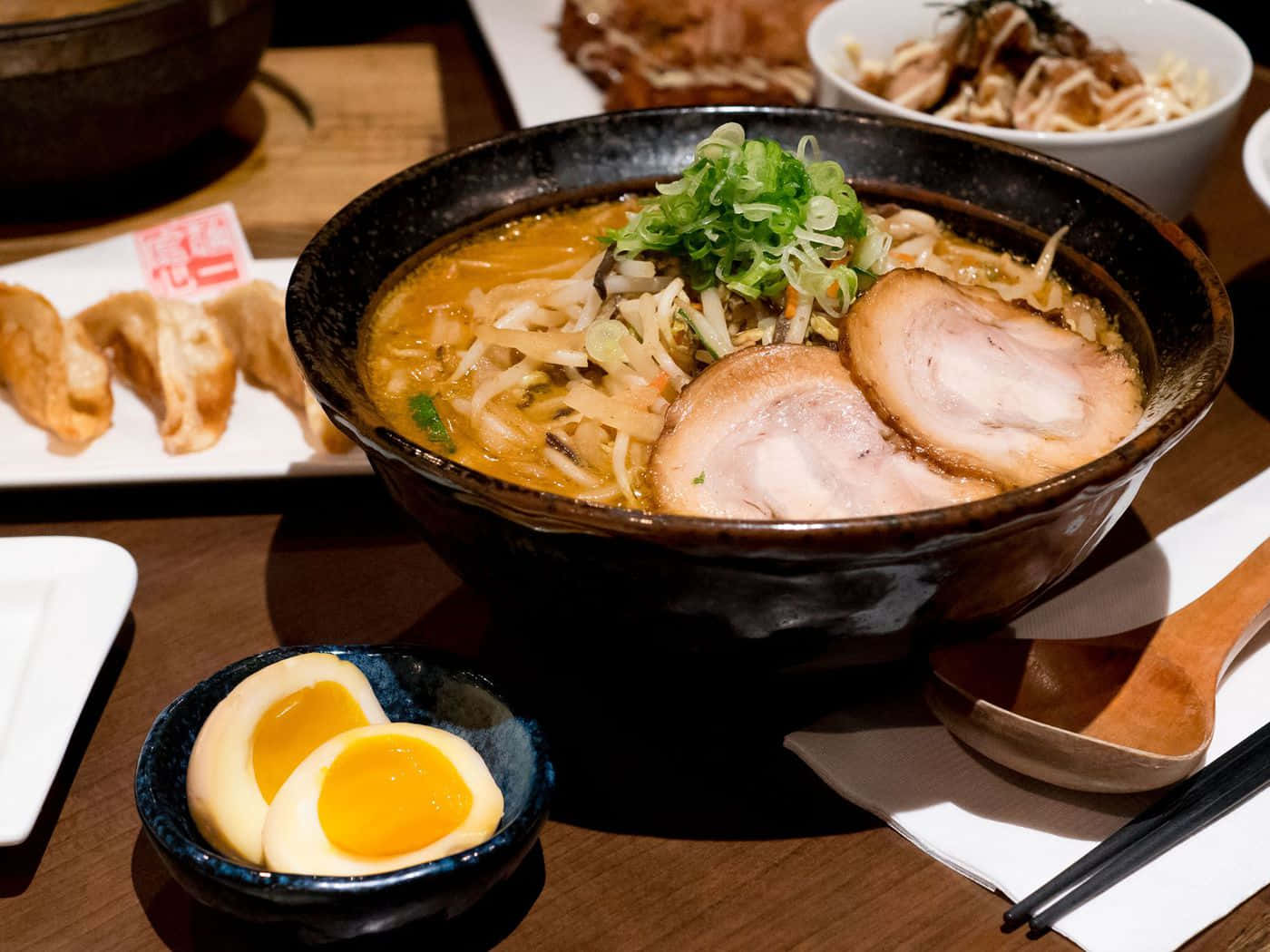 Noodles 桌布 壁紙 — 一碗熱騰騰的濃郁拉麵，配上嫩滑叉燒、溏心蛋和蔥花，放在木桌上，旁邊還有小菜。