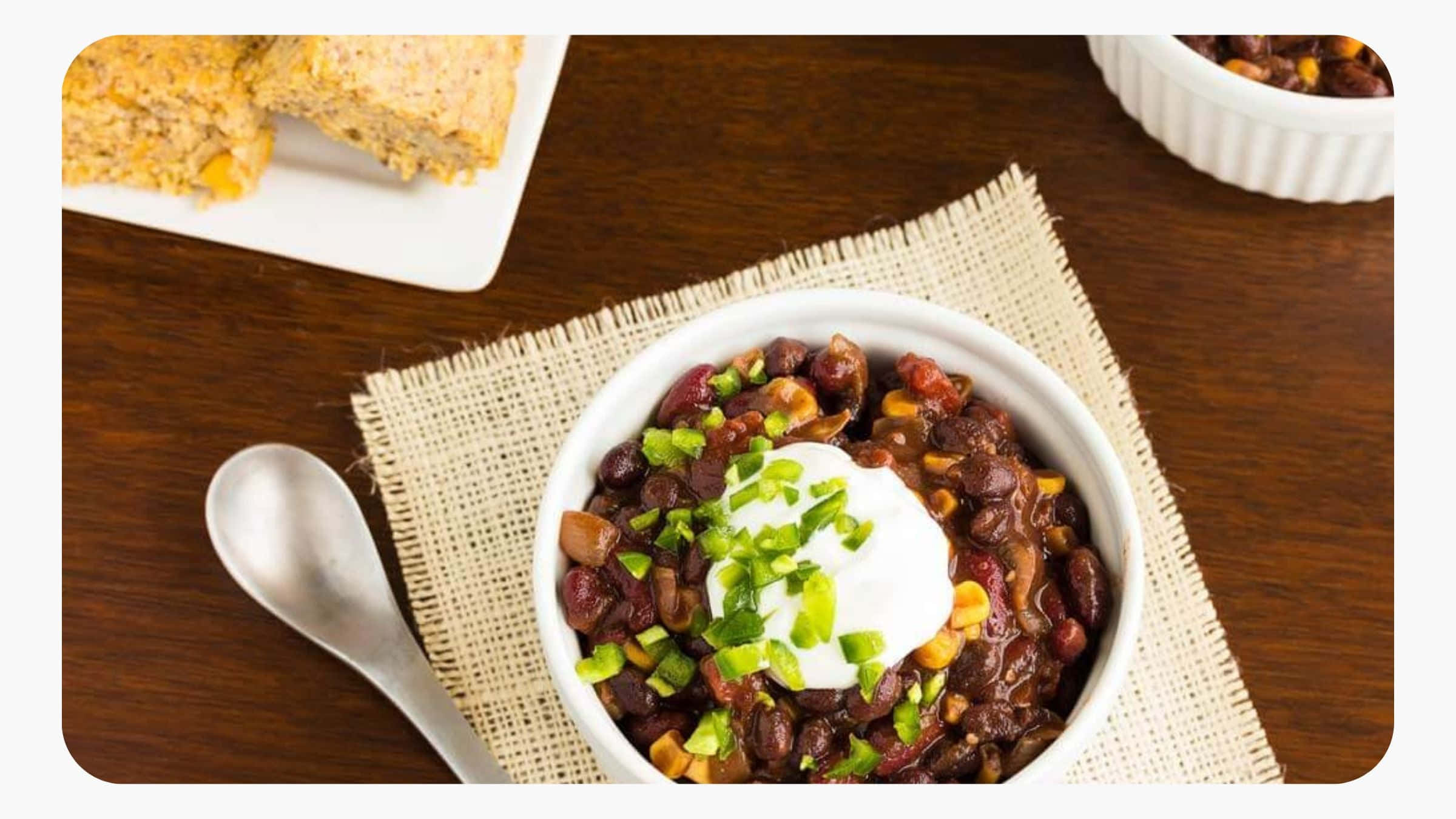 Black Bean Soup 桌布 壁紙 — 一碗熱騰騰的濃郁深色辣椒，上面放有酸奶油和切碎的青蔥，放在編織墊上，旁邊有玉米麵包和湯匙。