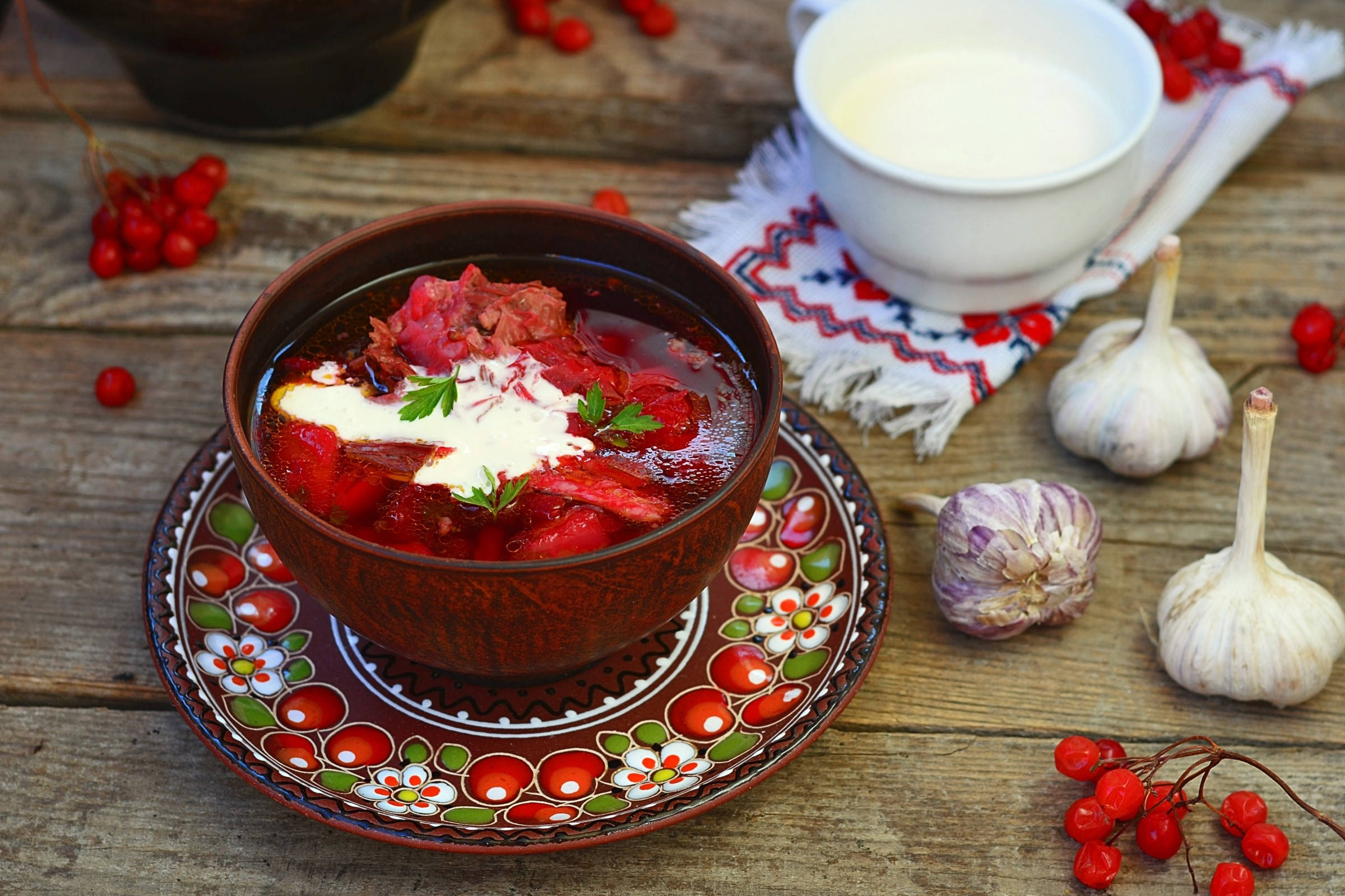 beetroot 桌布 壁紙 — 一碗熱騰騰的濃郁紅菜湯，配上酸奶油和歐芹，放在花卉圖案的盤子上，旁邊有大蒜和紅醋栗，置於鄉村風格的木桌上。